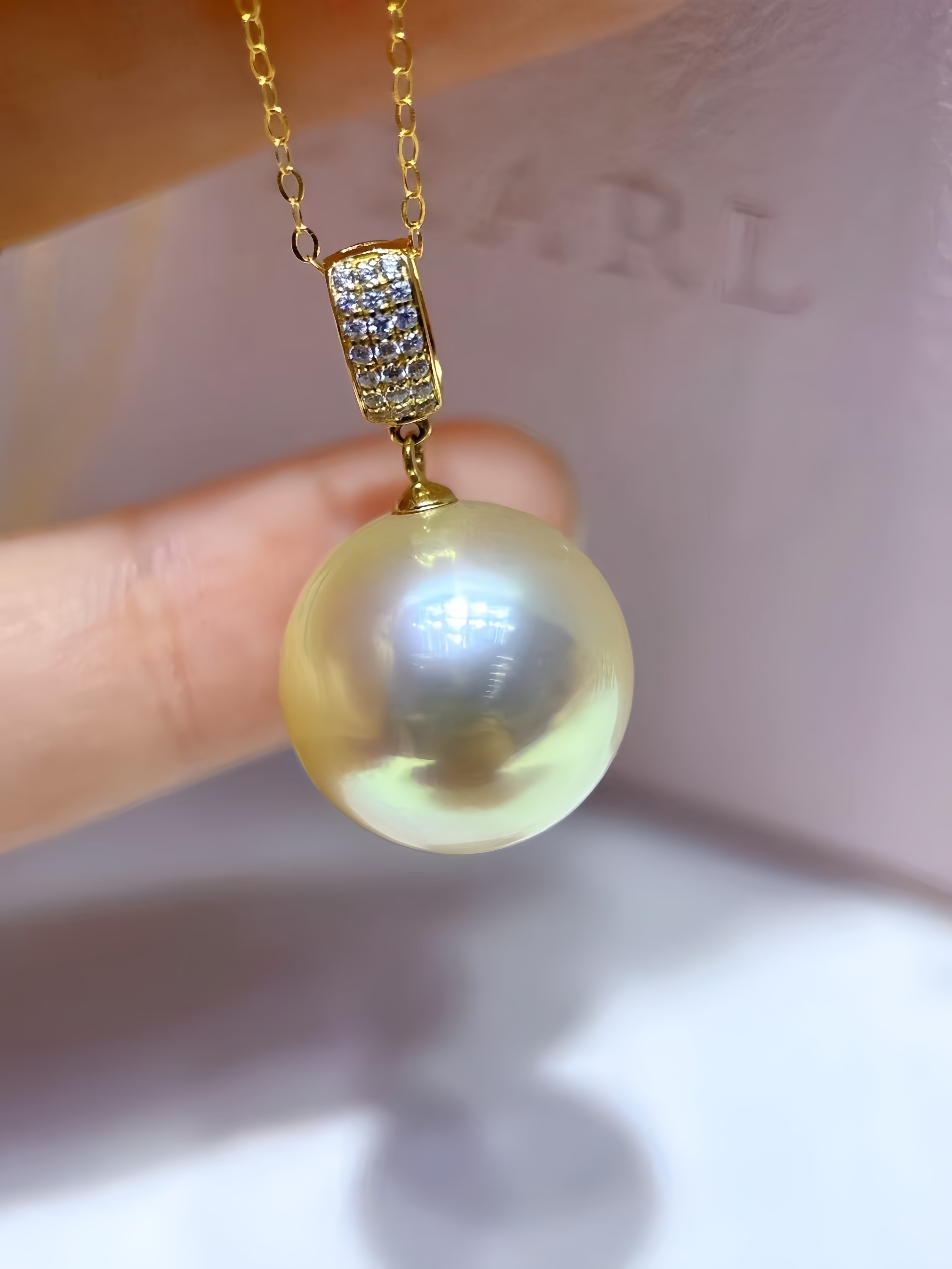 A close-up of a gold pendant necklace featuring a large, round, lustrous pearl pendant and a gold bail set with small clear stones, held by fingers against a soft pink background.