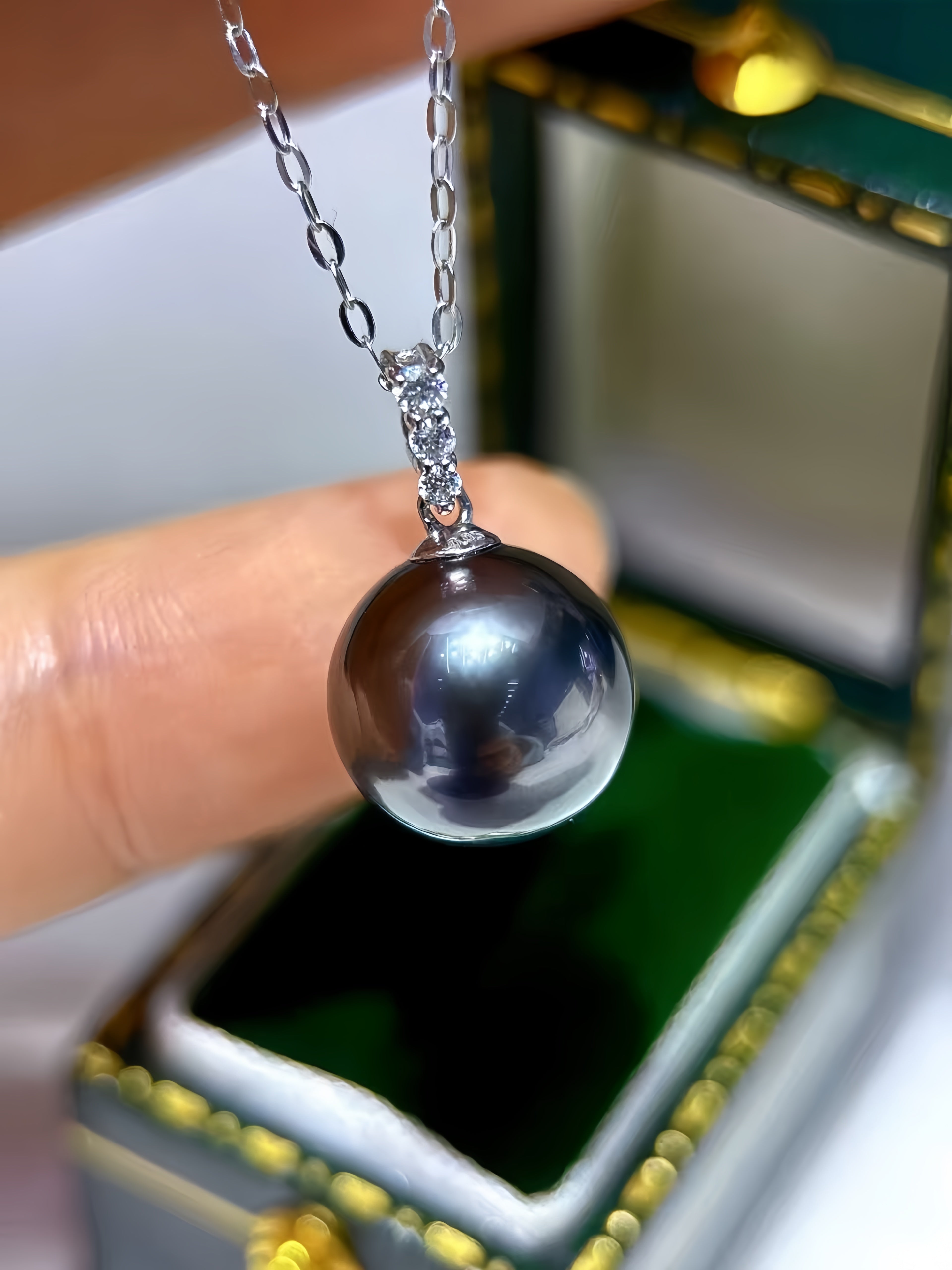 A close-up of a hand holding a silver pendant necklace with a large, dark, lustrous oval pearl. The necklace is positioned in front of an open green and gold jewelry box.