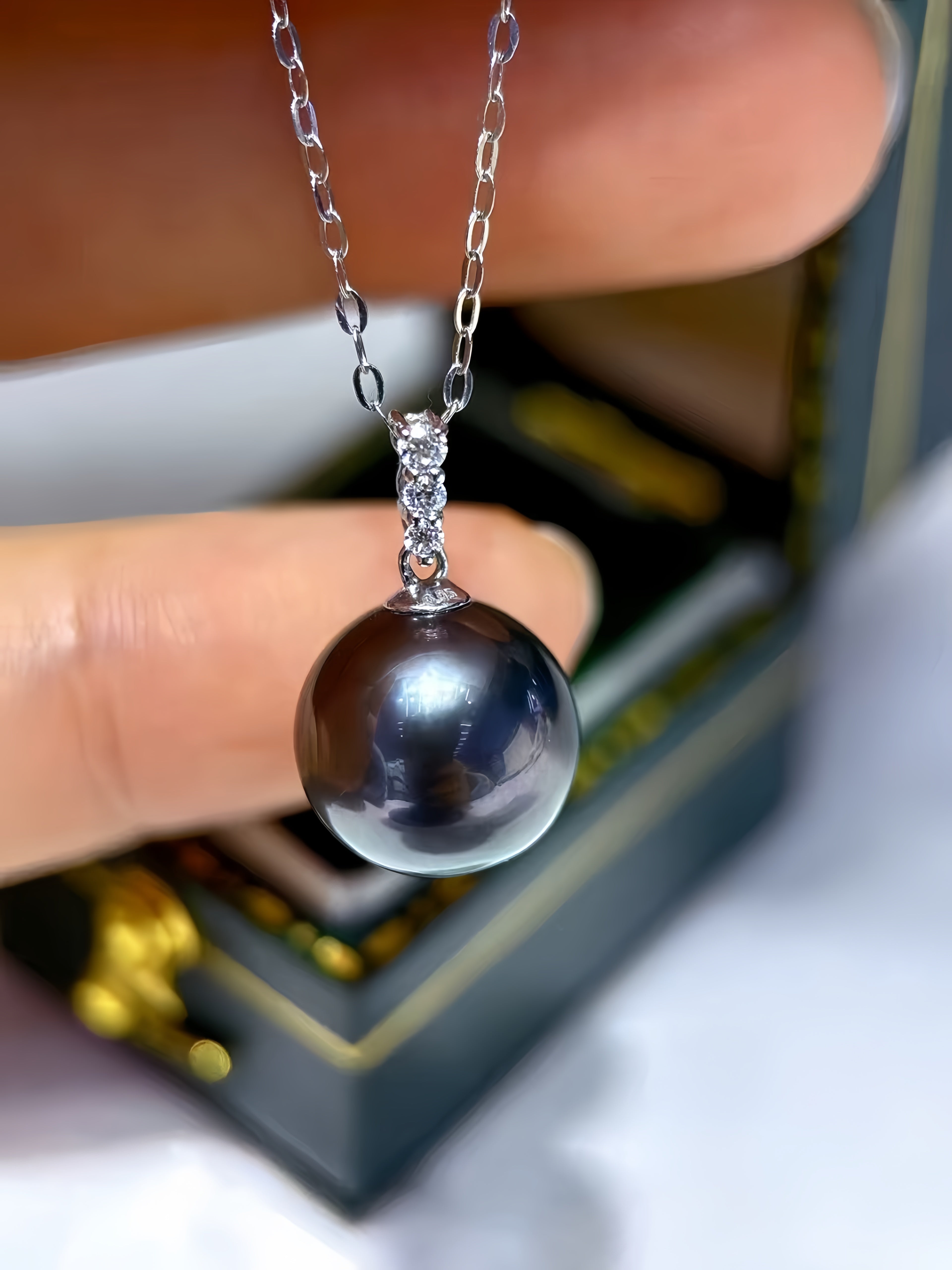 A close-up of a silver chain necklace with a large, dark, iridescent pearl pendant held by fingers, with a blurred jewelry box in the background.