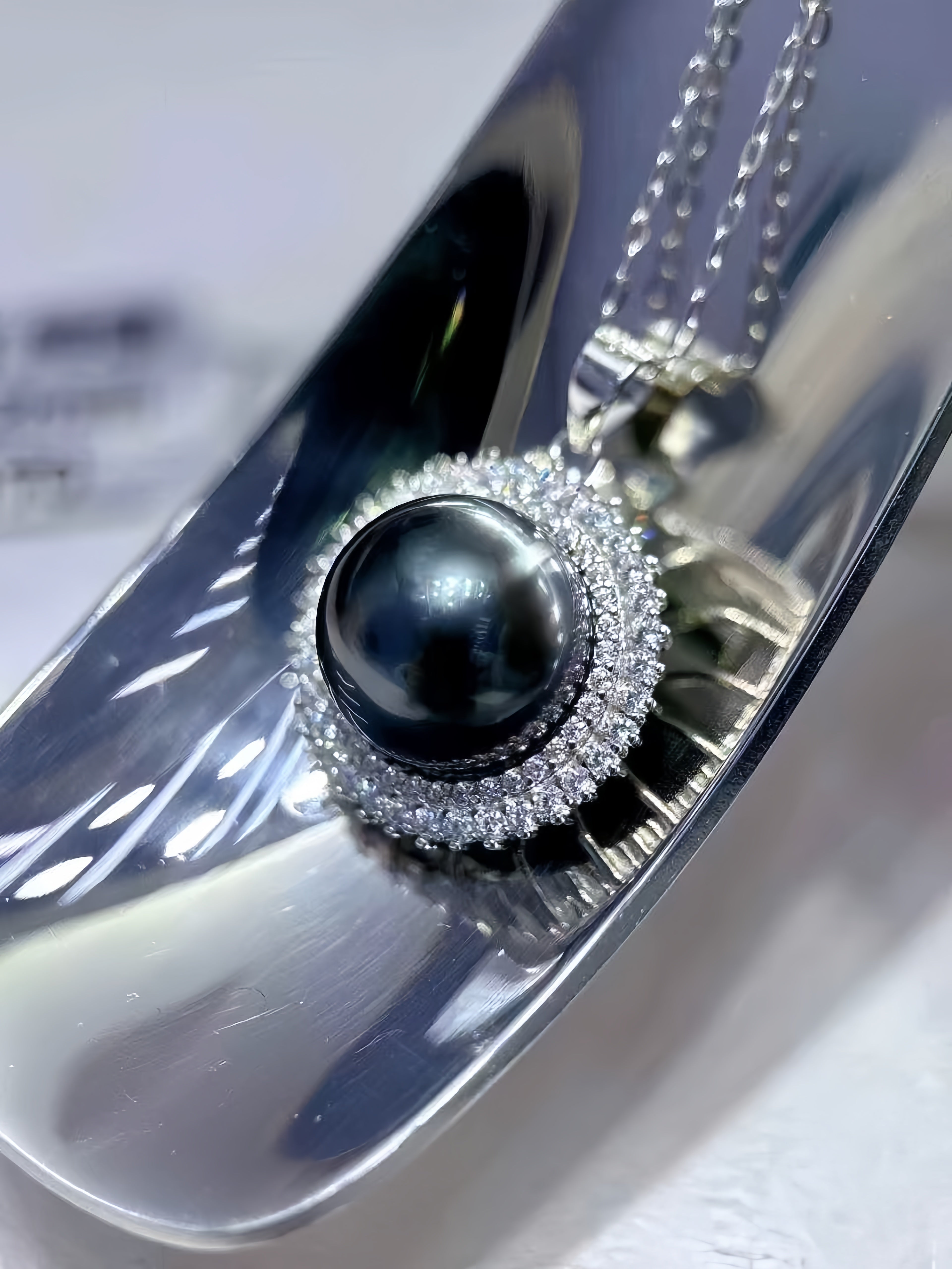 A close-up of a silver pendant necklace featuring a round black pearl surrounded by a halo of small clear stones, displayed on a reflective silver surface.