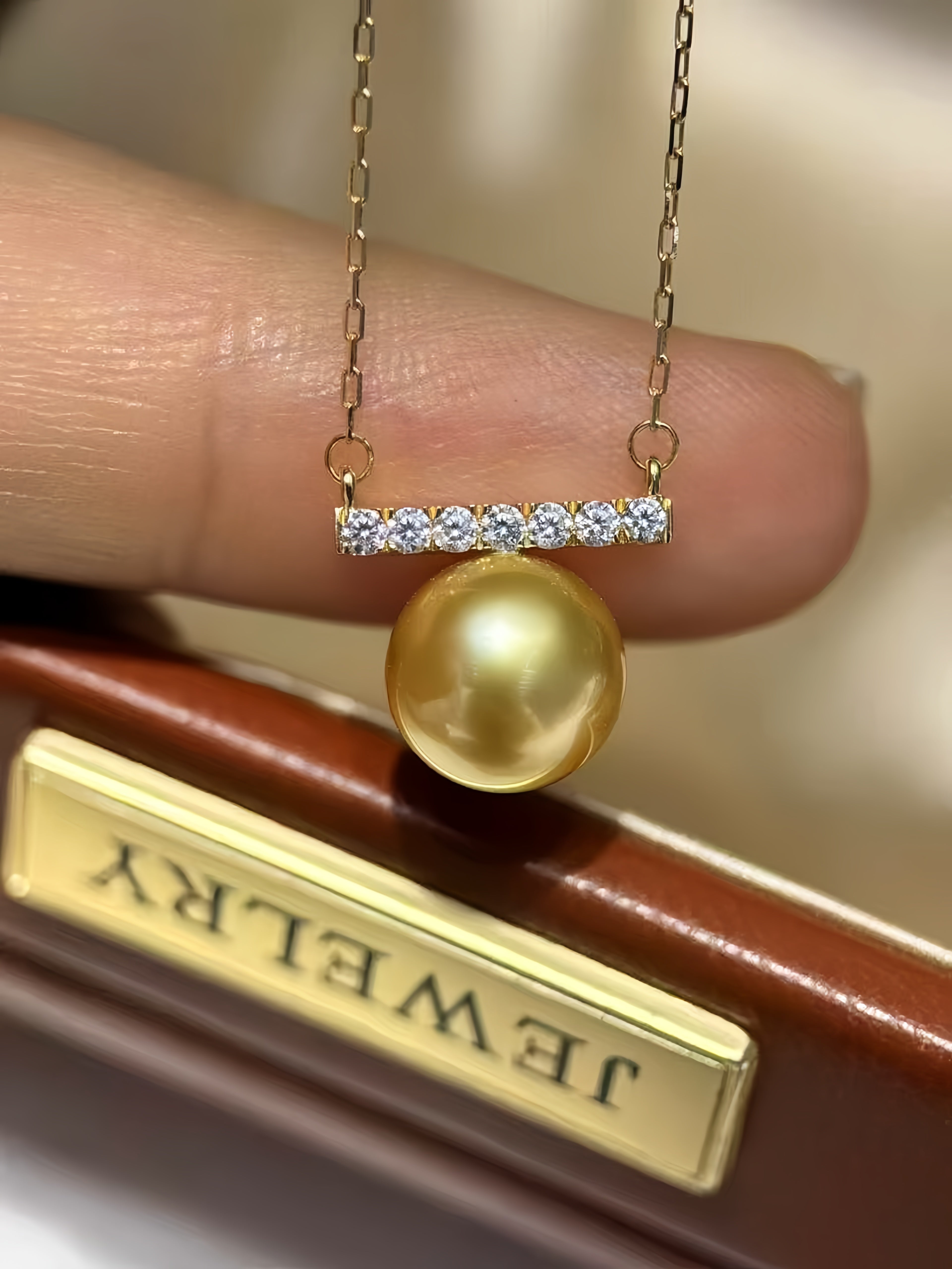 A gold necklace with a bar set with small clear stones and a single round gold pearl pendant, held against a finger with a jewelry box in the background.