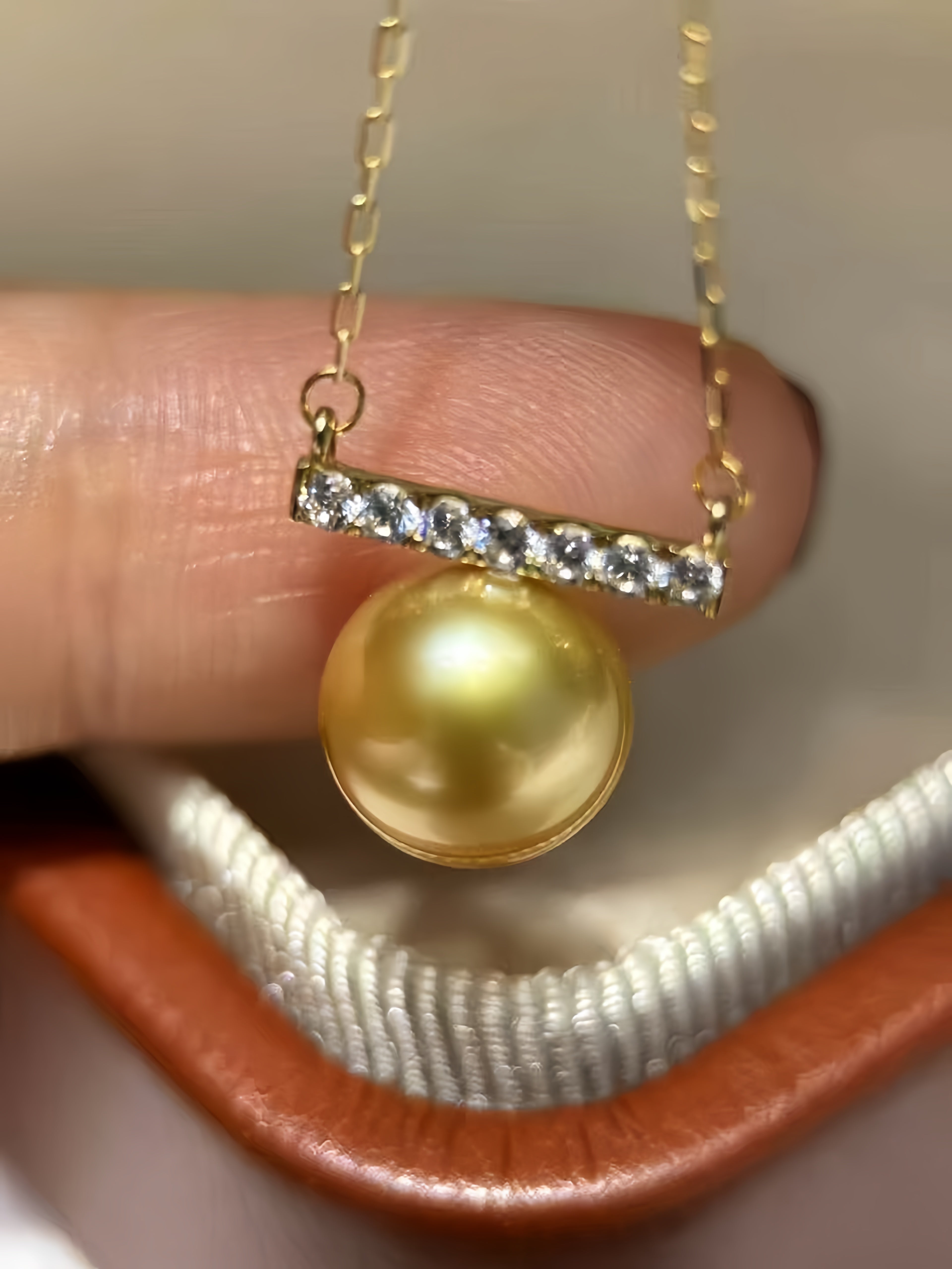 A gold pendant necklace featuring a round yellow pearl and a bar-set clear stone accent, held between fingers over a white and brown jewelry box.
