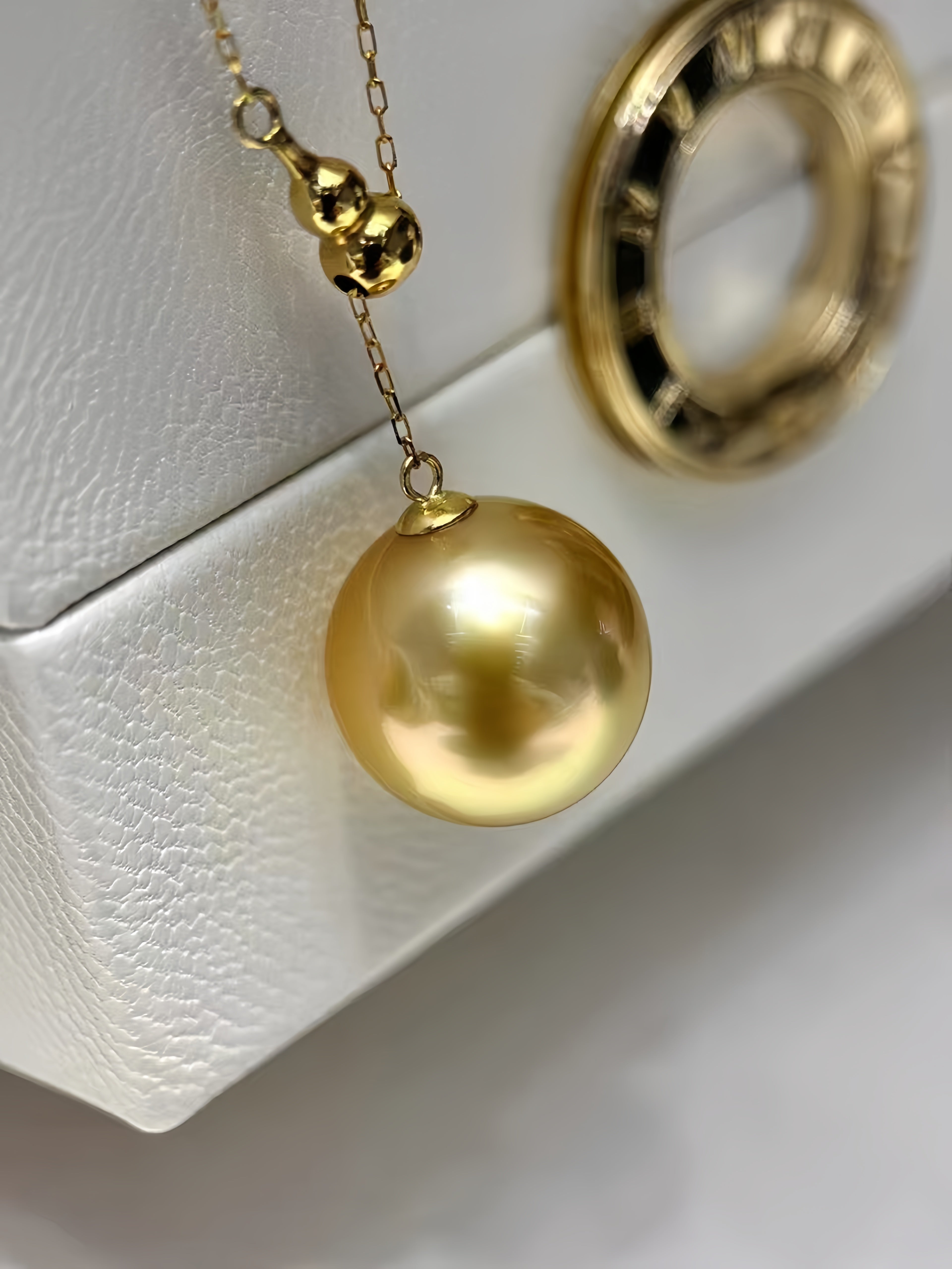 A gold pendant necklace featuring a large, lustrous gold pearl and a small gold gourd charm, displayed on a textured white surface.