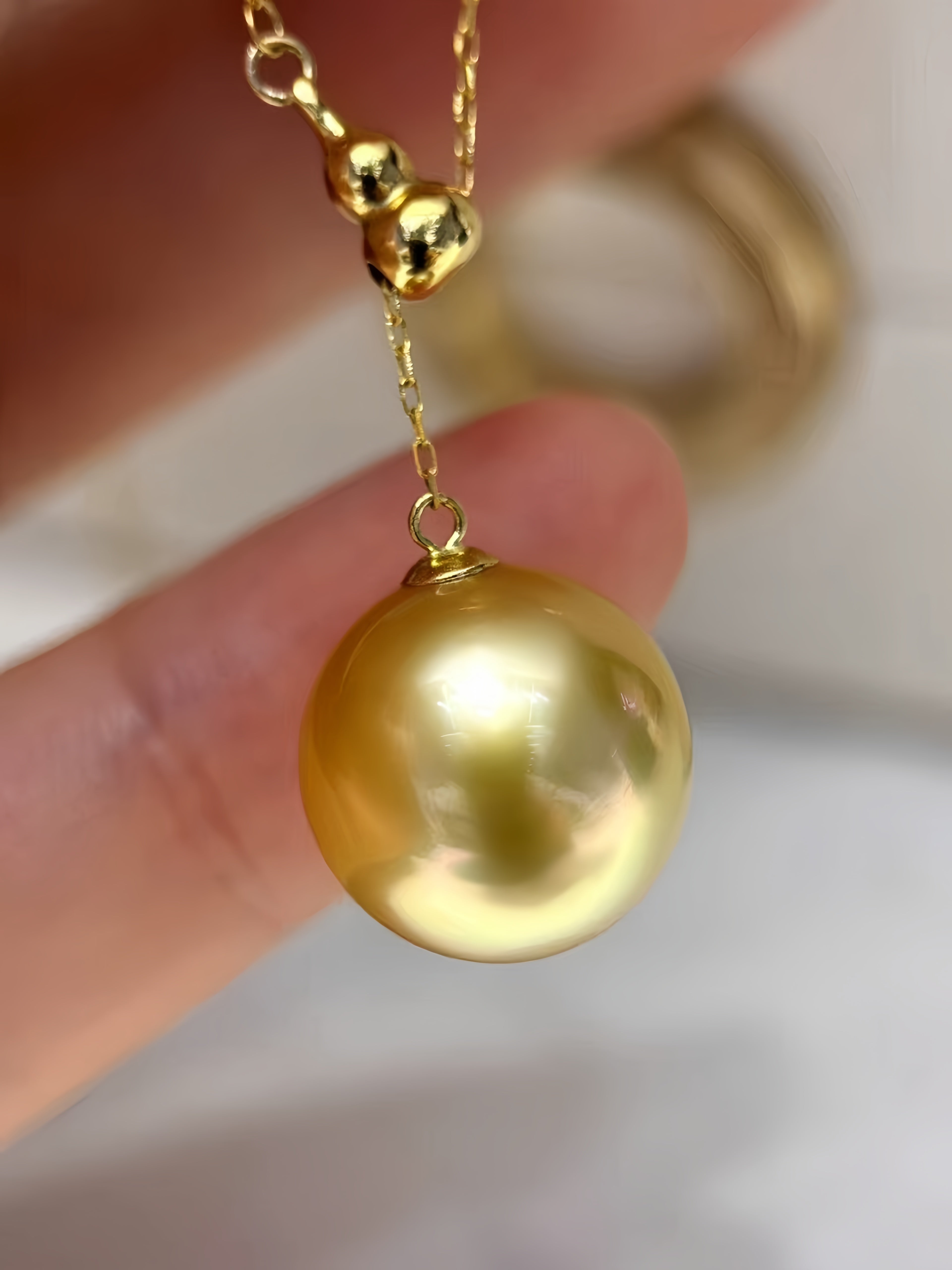 A close-up of a gold necklace with a large, lustrous gold pearl pendant held between fingers, with a blurred gold ring in the background.