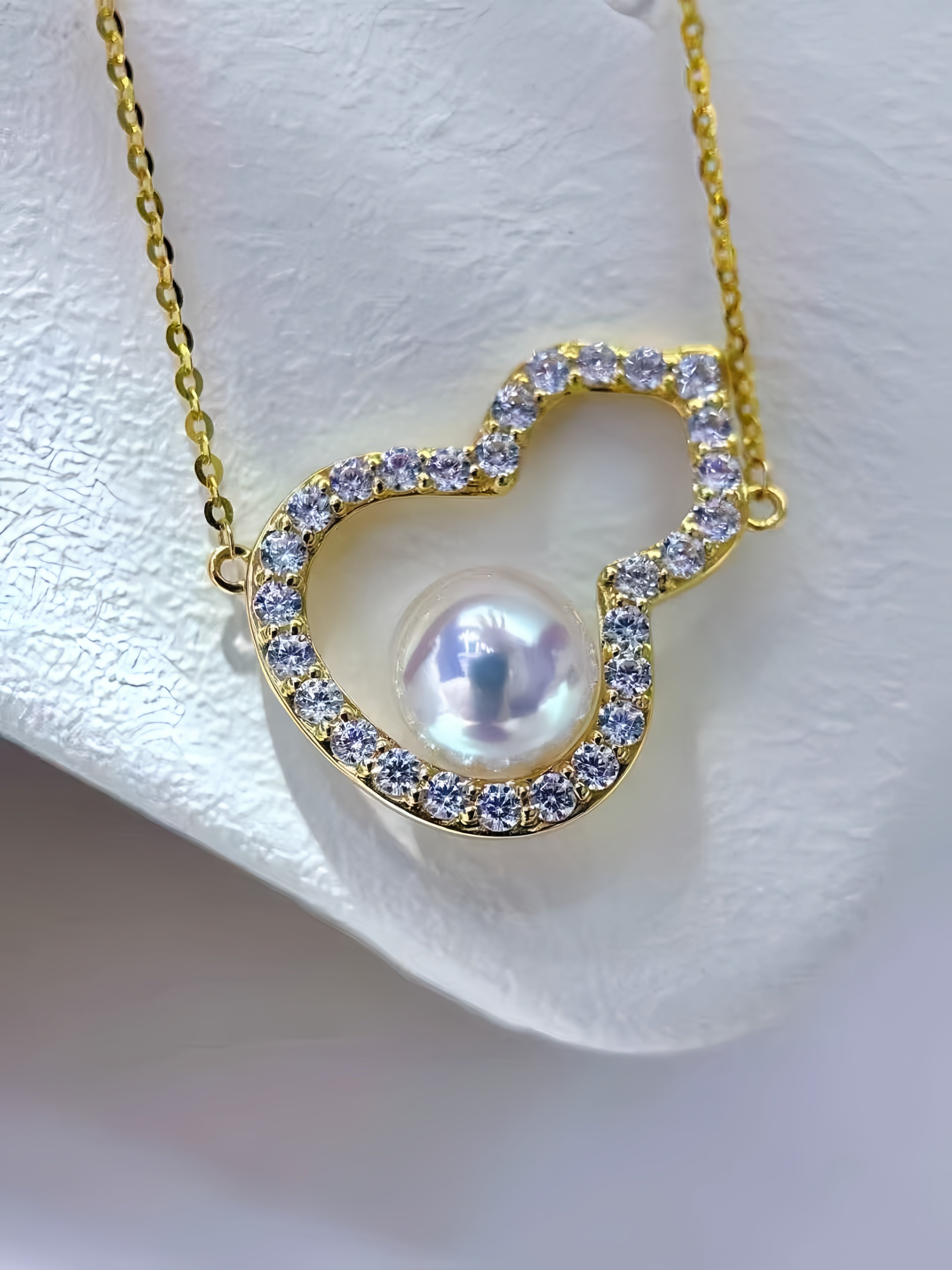 A gold heart-shaped pendant necklace featuring a central lustrous pearl surrounded by small clear stones, displayed on a white textured stand.