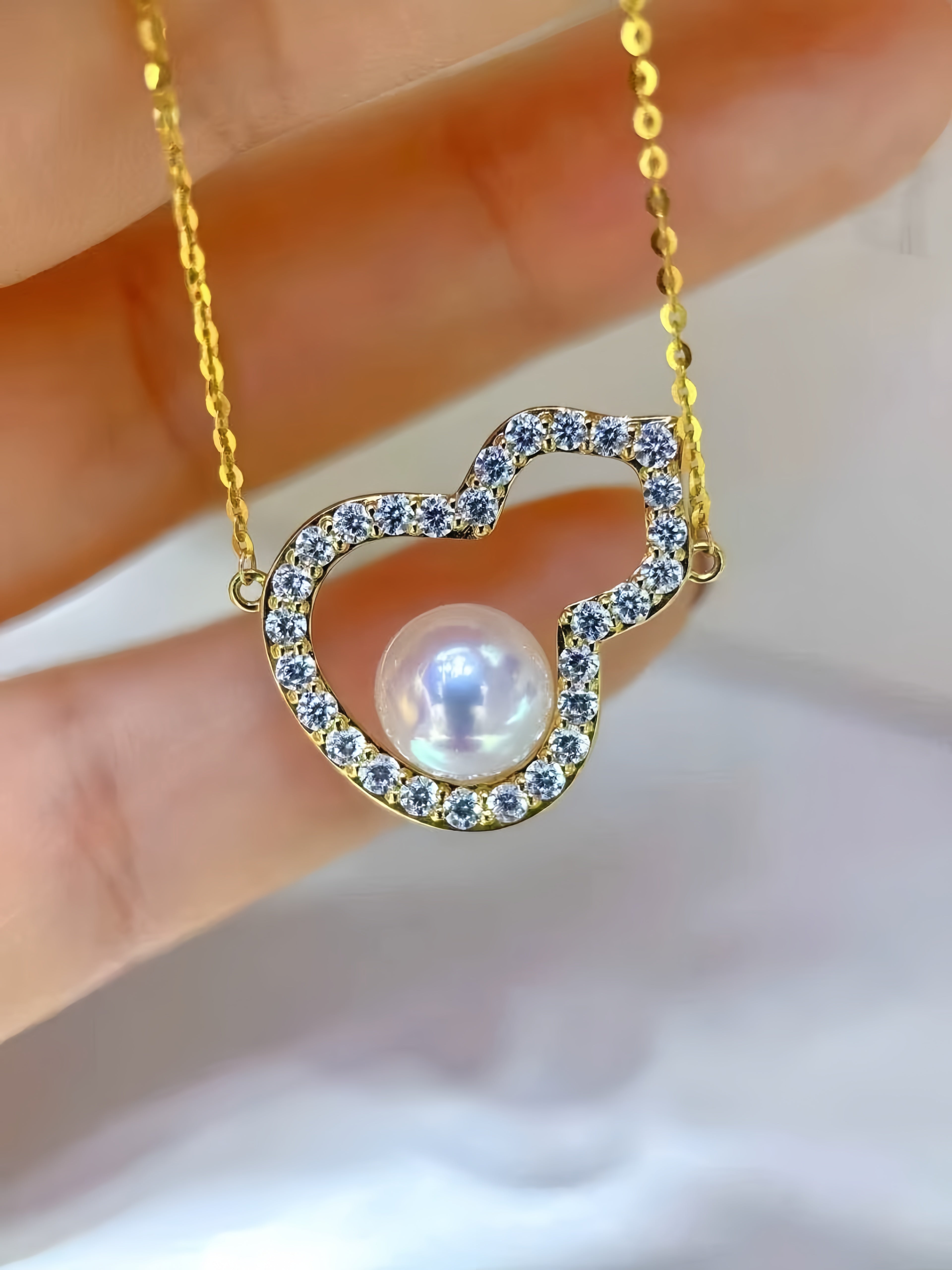 A gold heart-shaped pendant necklace with a central white pearl, held by a hand. The heart outline is adorned with small blue stones, and the chain is gold.