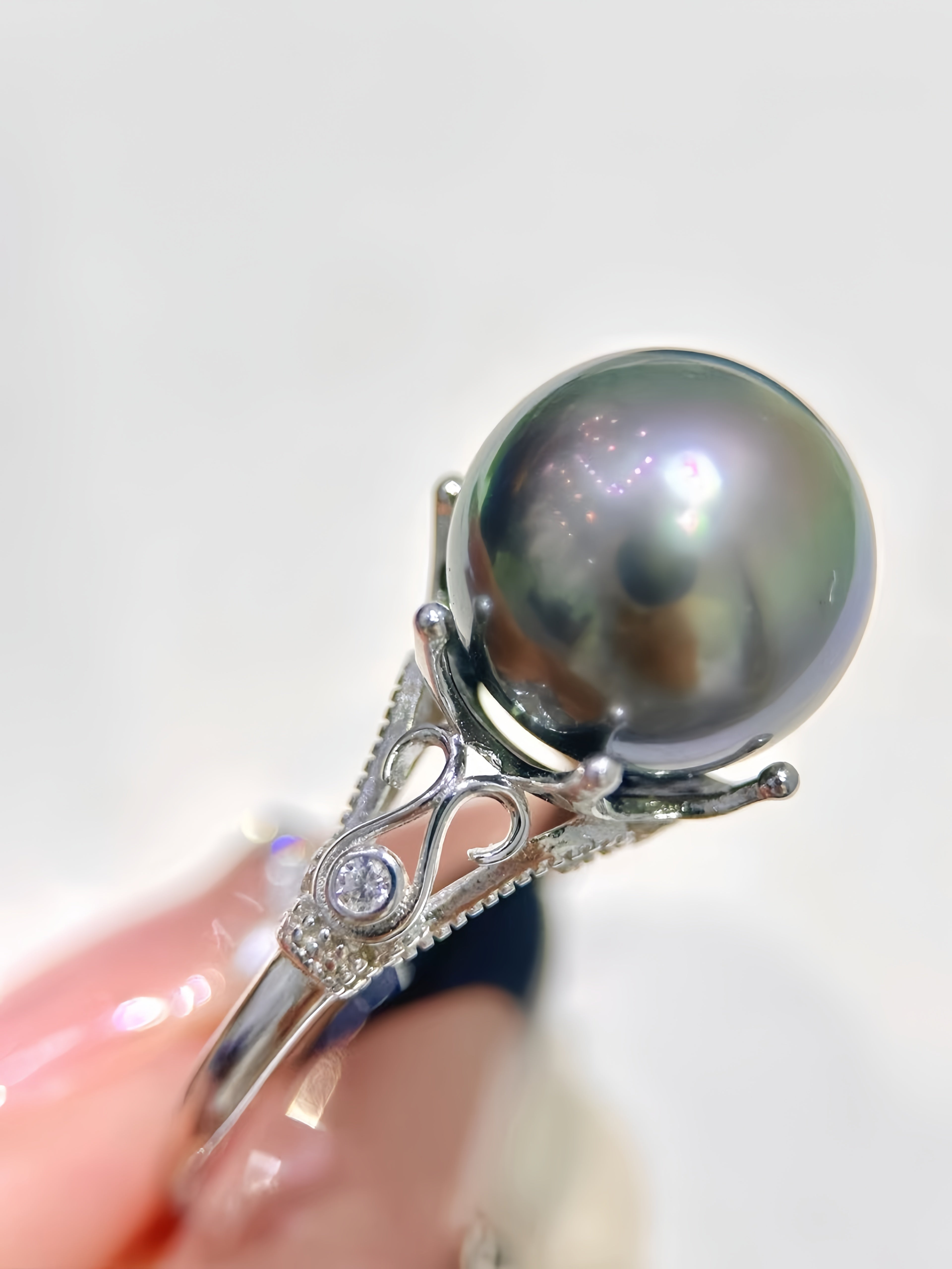 A close-up of a ring held between fingers, featuring a large round dark pearl on an ornate silver-toned setting with small clear stones, against a soft white background.