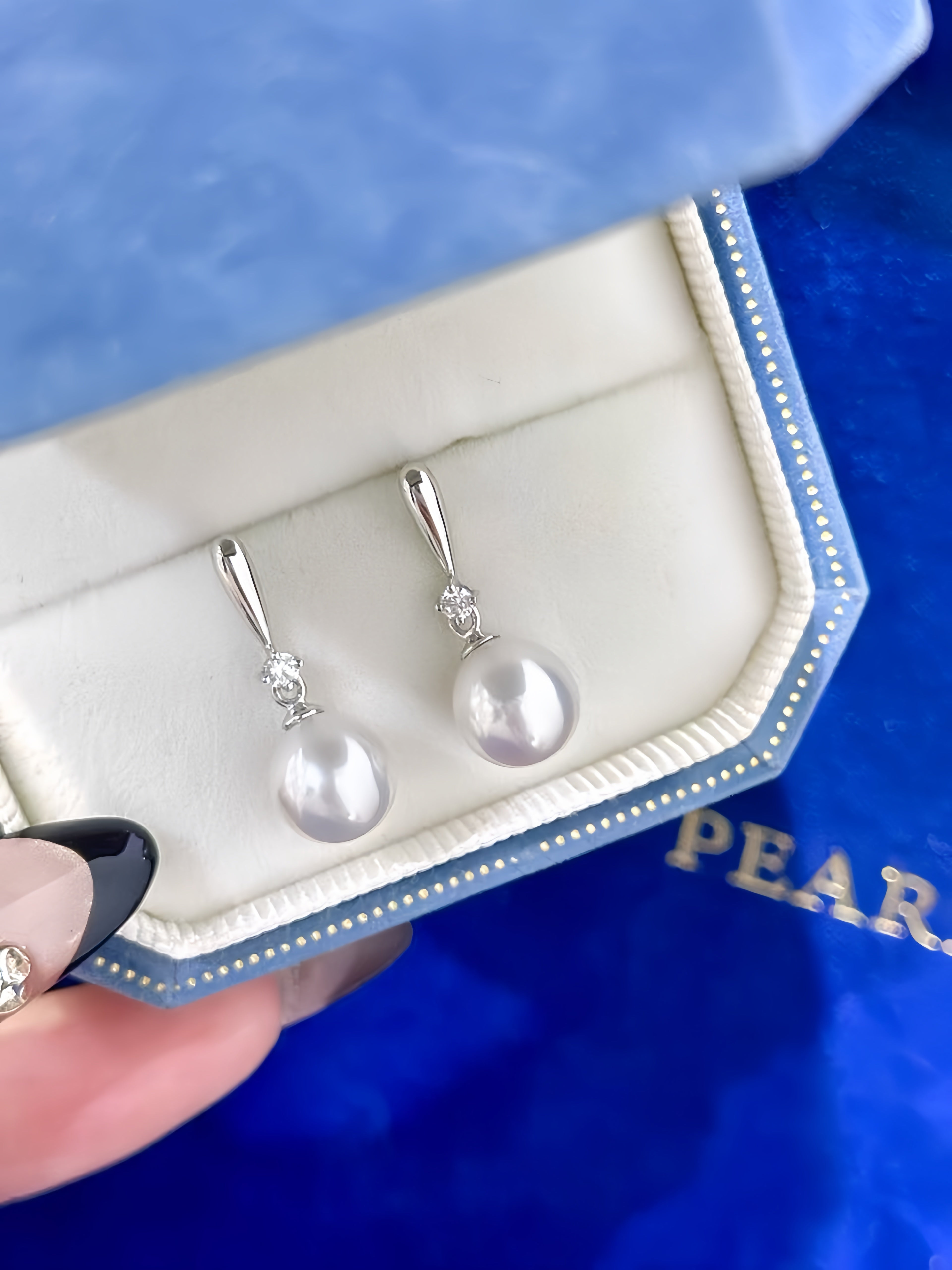 A pair of silver drop earrings with white freshwater pearls and small clear stones, displayed in a blue and white Pearls box held by a hand.