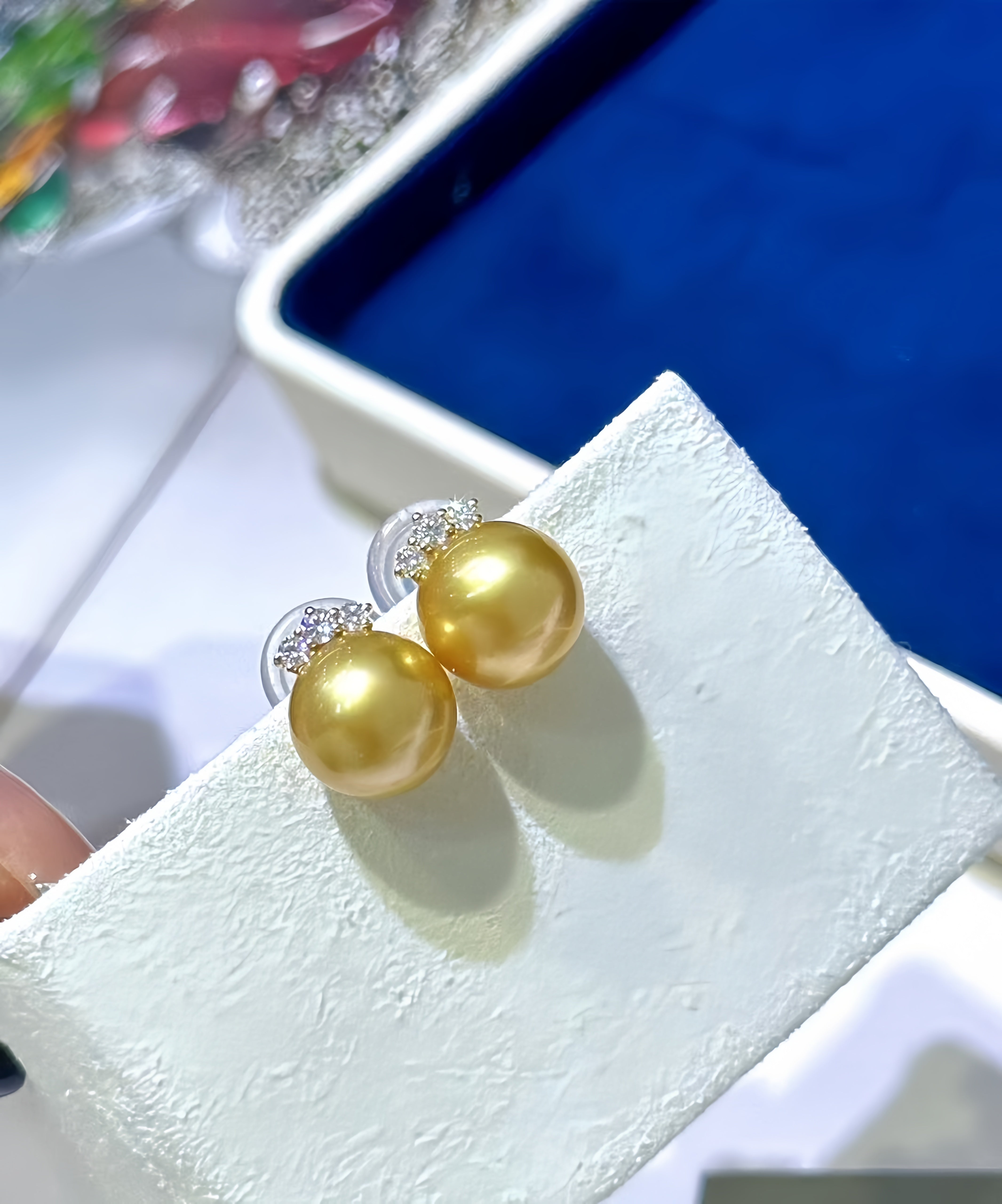 A pair of gold round pearl stud earrings with silver posts and clear stones, displayed on a white textured jewelry stand.