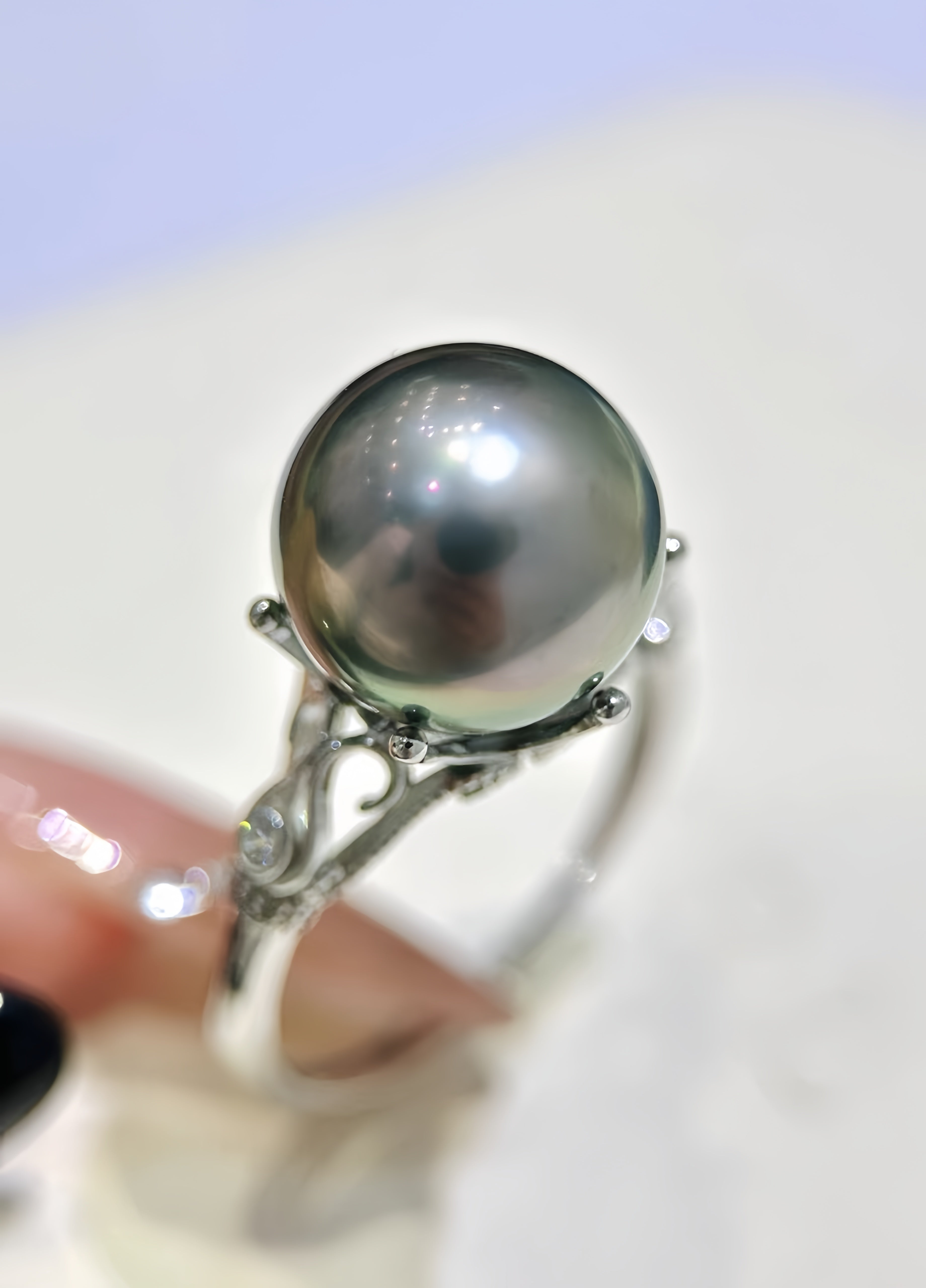 A silver ring featuring a large, round, dark gray pearl in a prong setting, held by a hand with manicured nails against a soft-focus light background.
