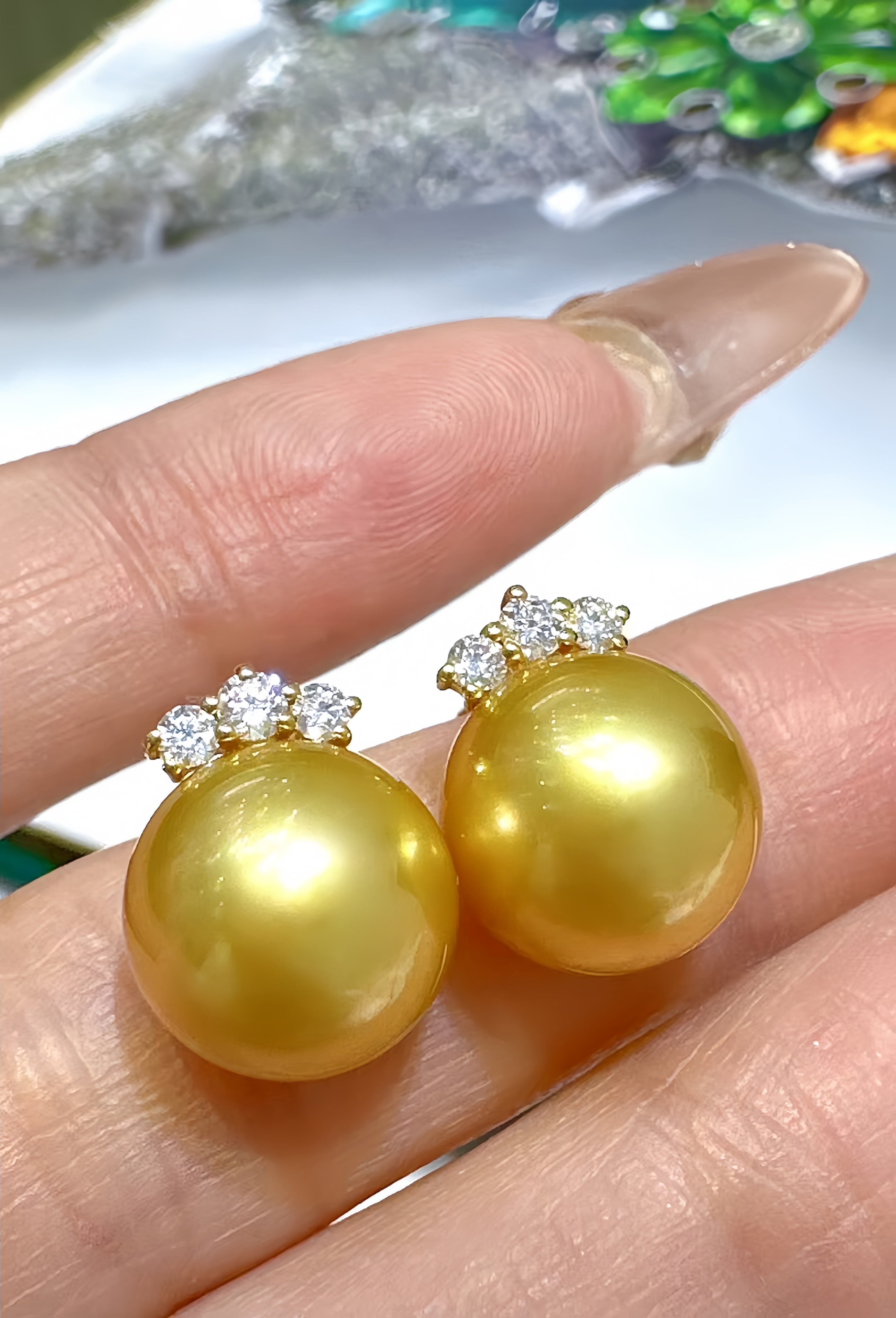 A pair of gold pearl stud earrings with small clear crystal clusters at the top, shown resting on a person's fingers.