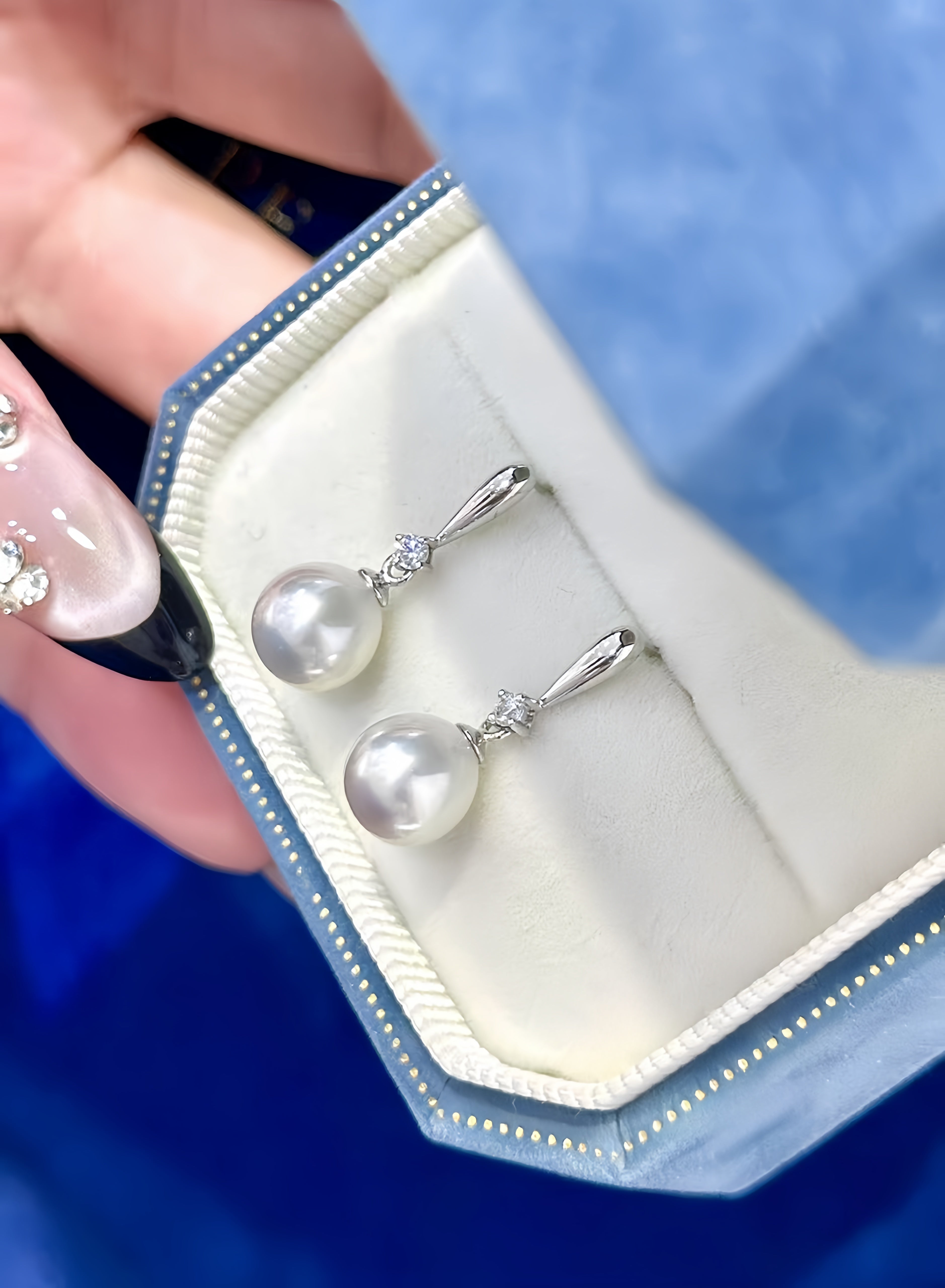 A pair of silver drop earrings with large white pearl-like beads and small clear stones, displayed in an open jewelry box held by a hand.