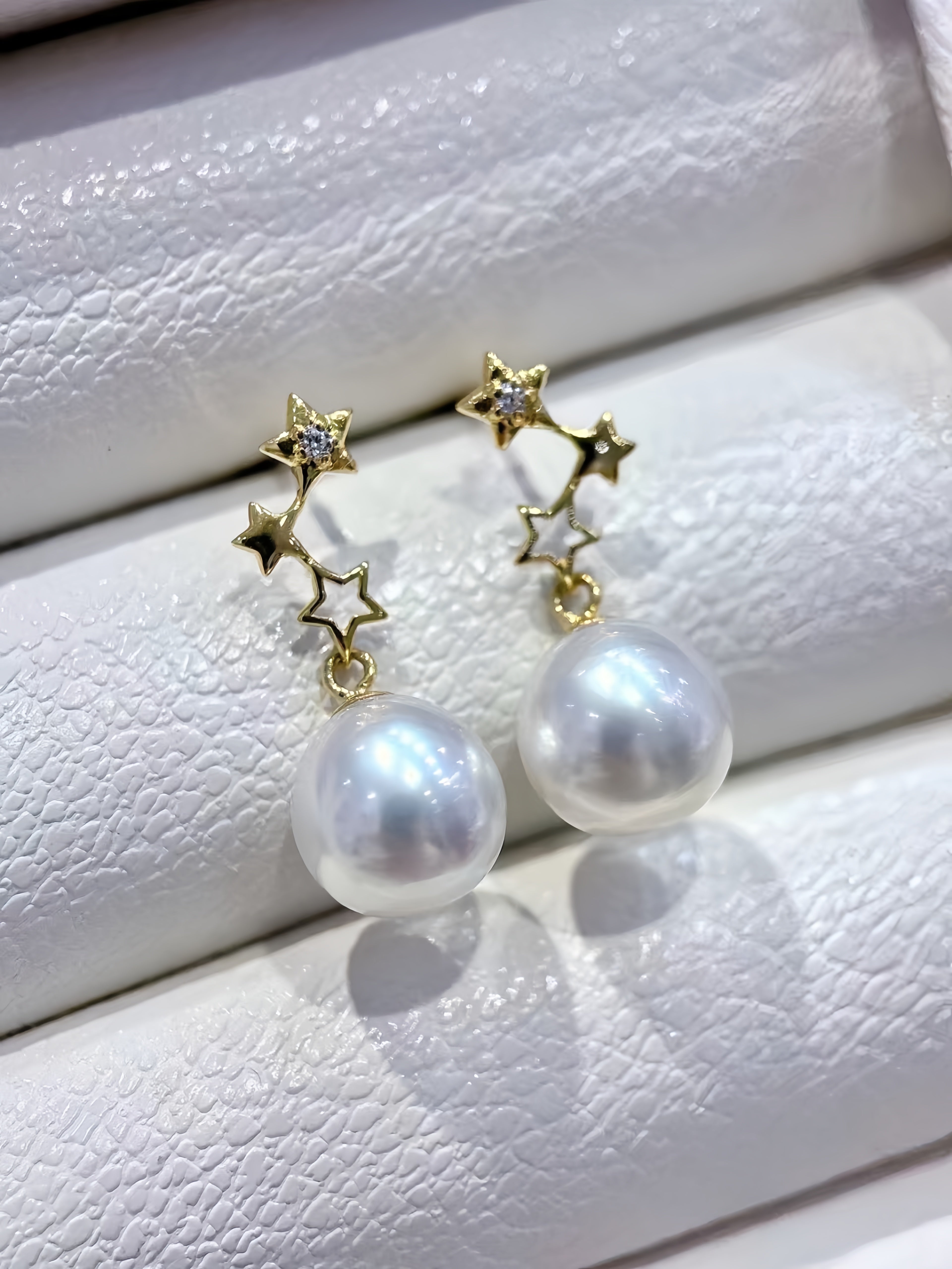 A pair of gold star-shaped dangle earrings with white freshwater pearls, displayed on a textured white jewelry stand.