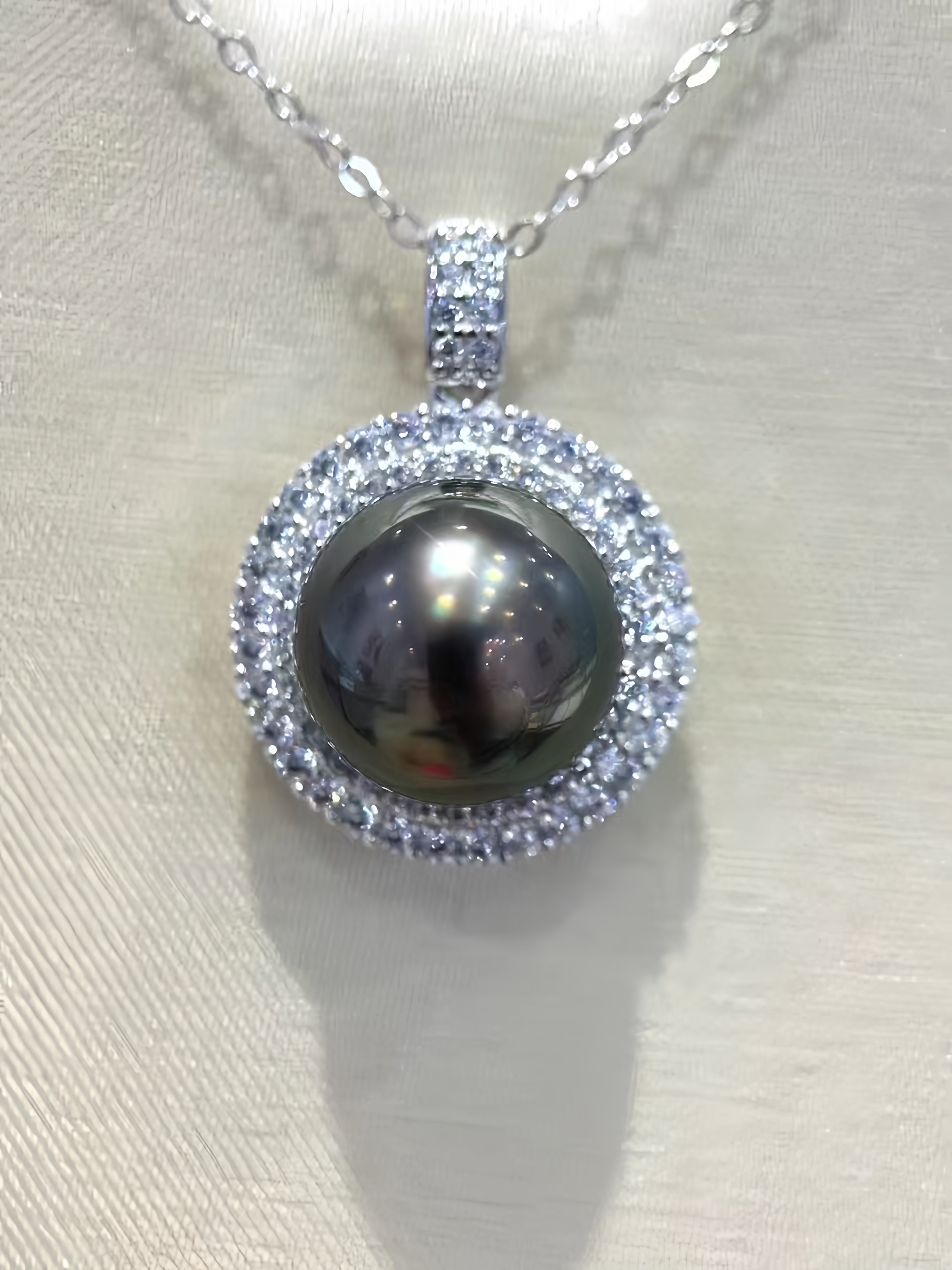 A silver pendant necklace featuring a large, dark, lustrous oval pearl surrounded by a halo of small clear stones, suspended from a silver chain on a light beige textured fabric background.