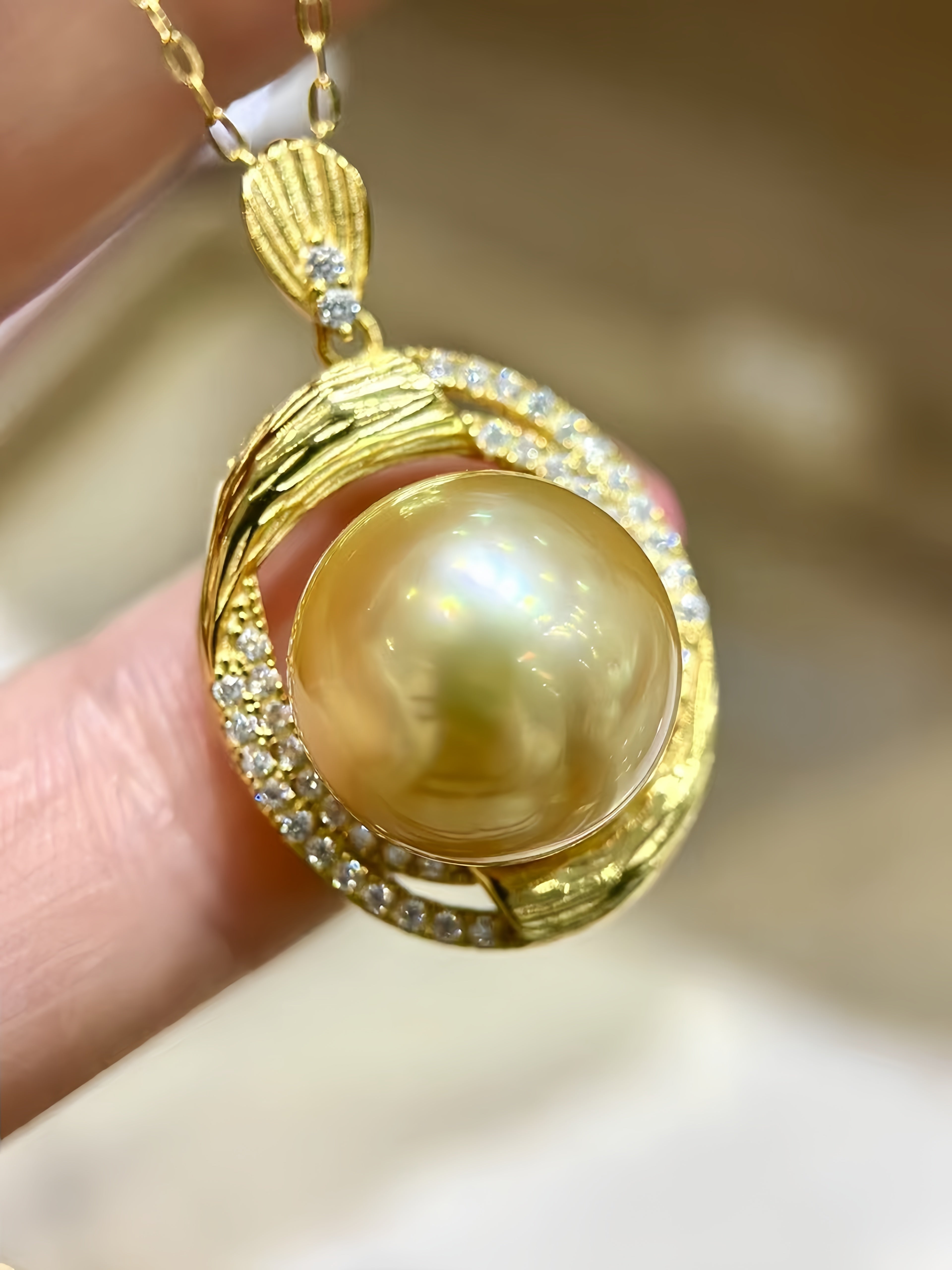 A close-up of a gold pendant necklace featuring a large round yellow pearl surrounded by small clear stones, held between fingers against a softly blurred beige background.