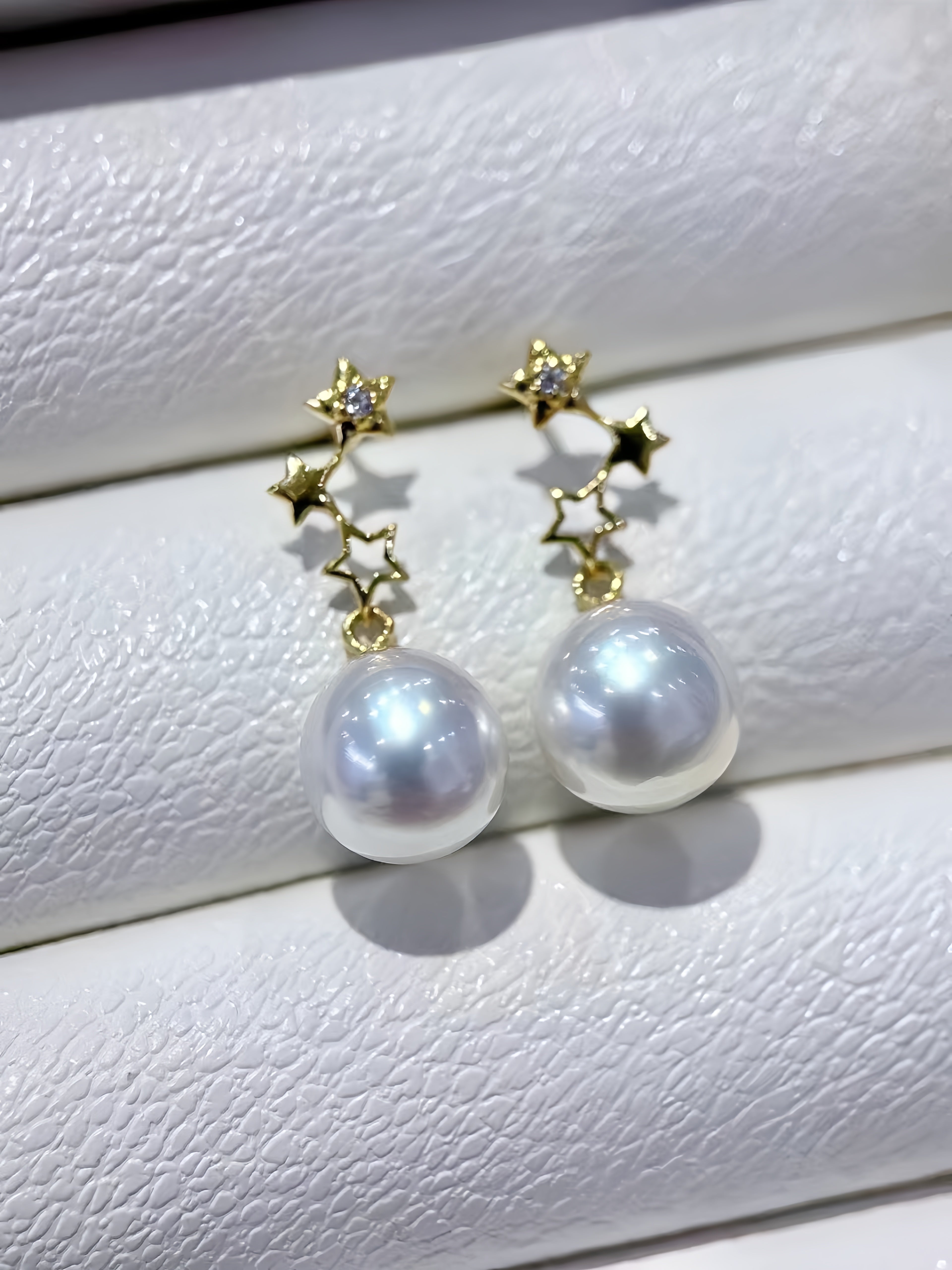 A pair of gold star-shaped dangle earrings with large white freshwater pearls, displayed on a textured white surface.