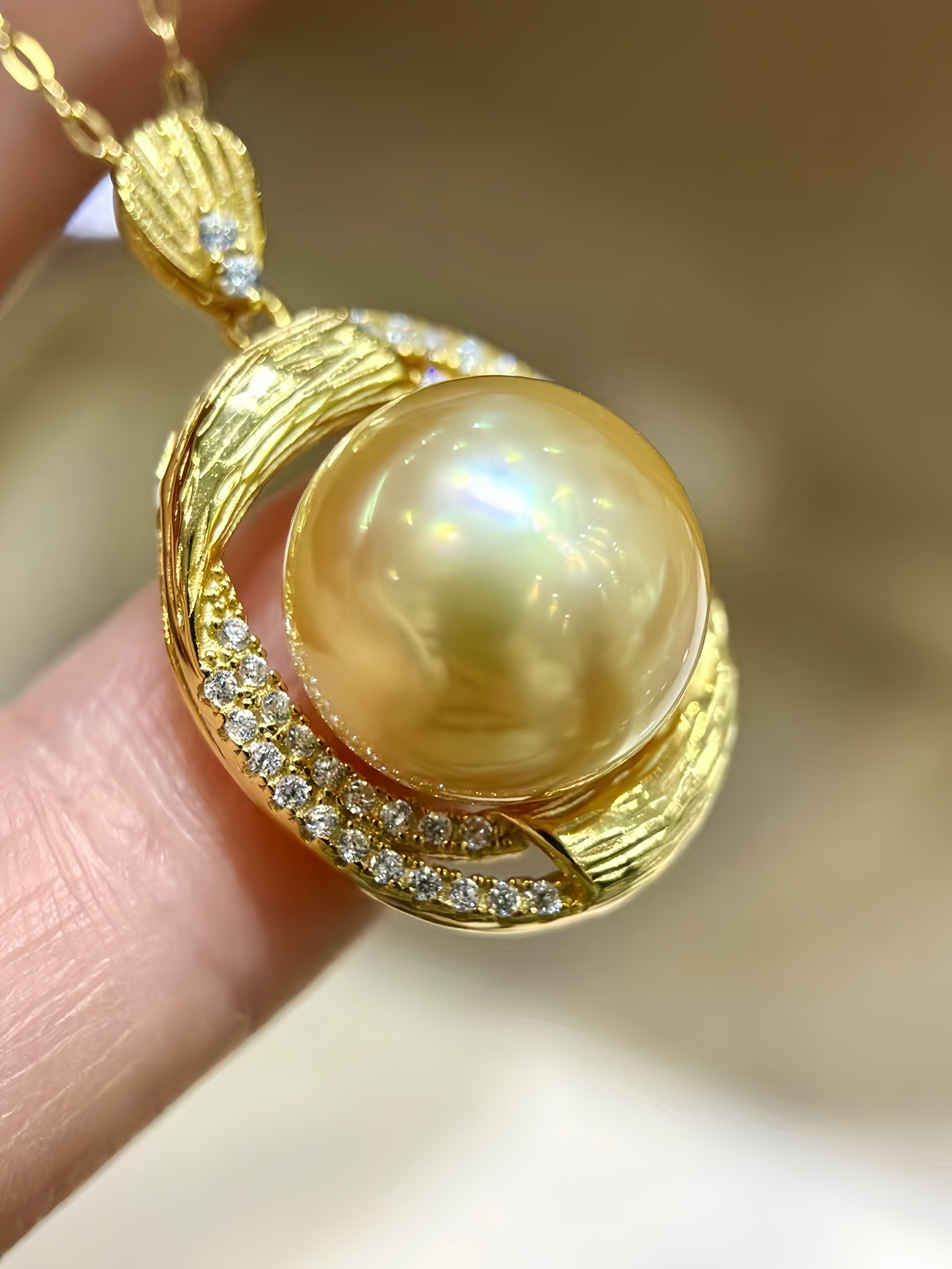 A close-up of a gold pendant necklace featuring a large round iridescent pearl surrounded by gold curved bands and small clear stones, held between fingers against a soft blurred beige background.