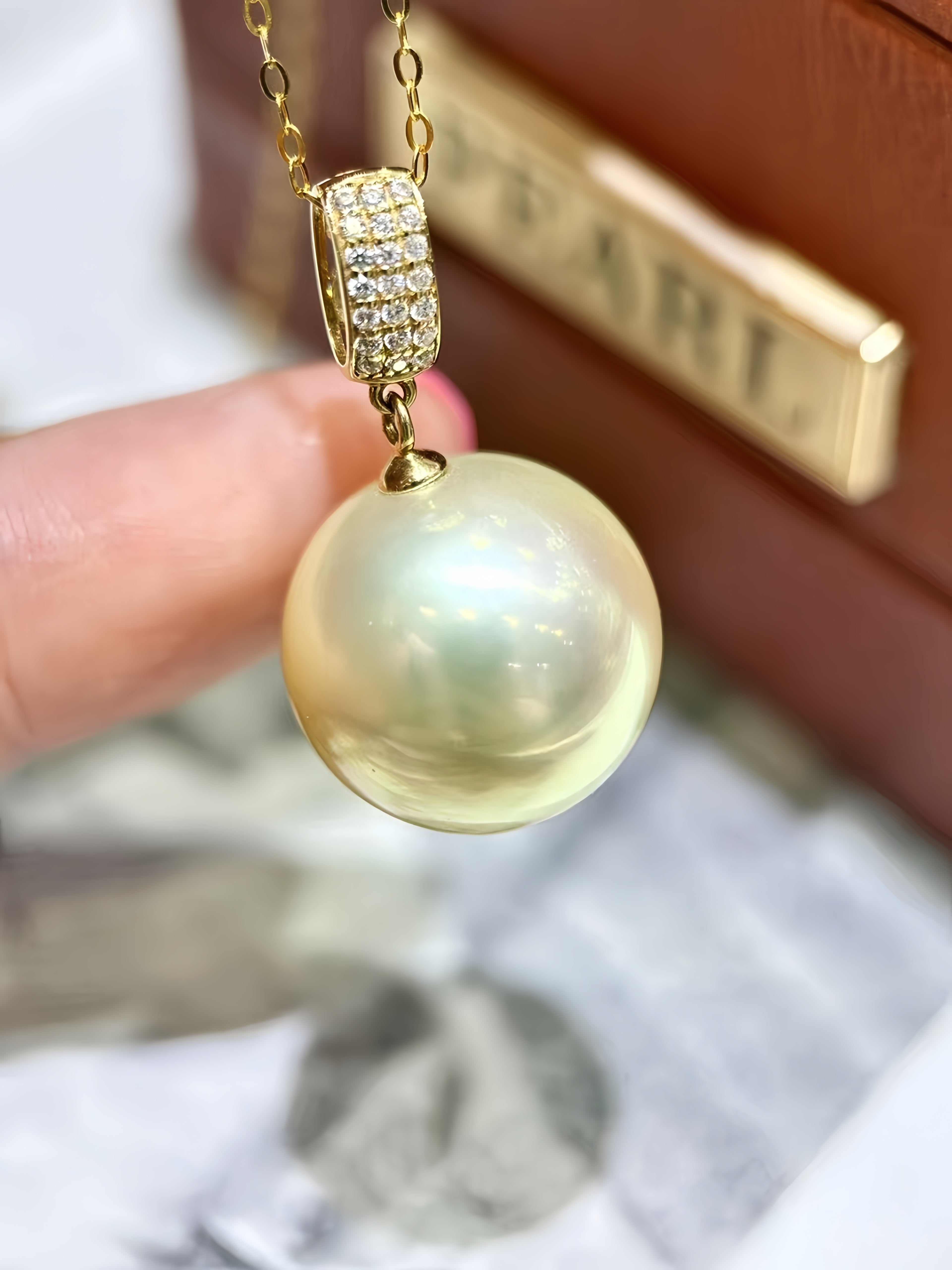 A gold pendant necklace featuring a large, lustrous cream-colored pearl pendant and a gold clasp adorned with small clear stones, held by a hand in front of a blurred brown box with a gold brand plaque.