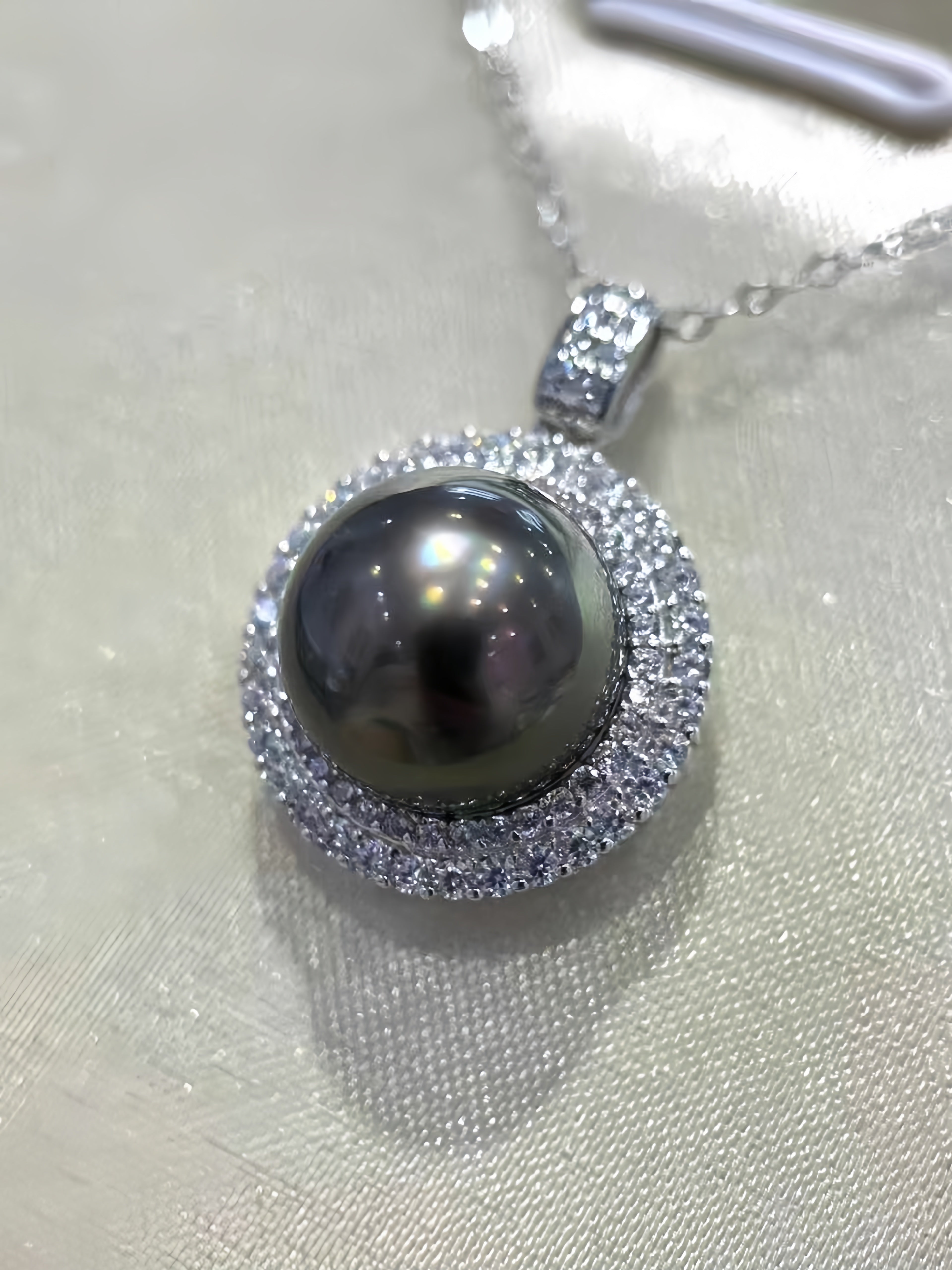 A close-up of a pendant necklace featuring a large round dark gray pearl surrounded by a halo of small clear stones, on a silver-toned chain against a light textured fabric background.
