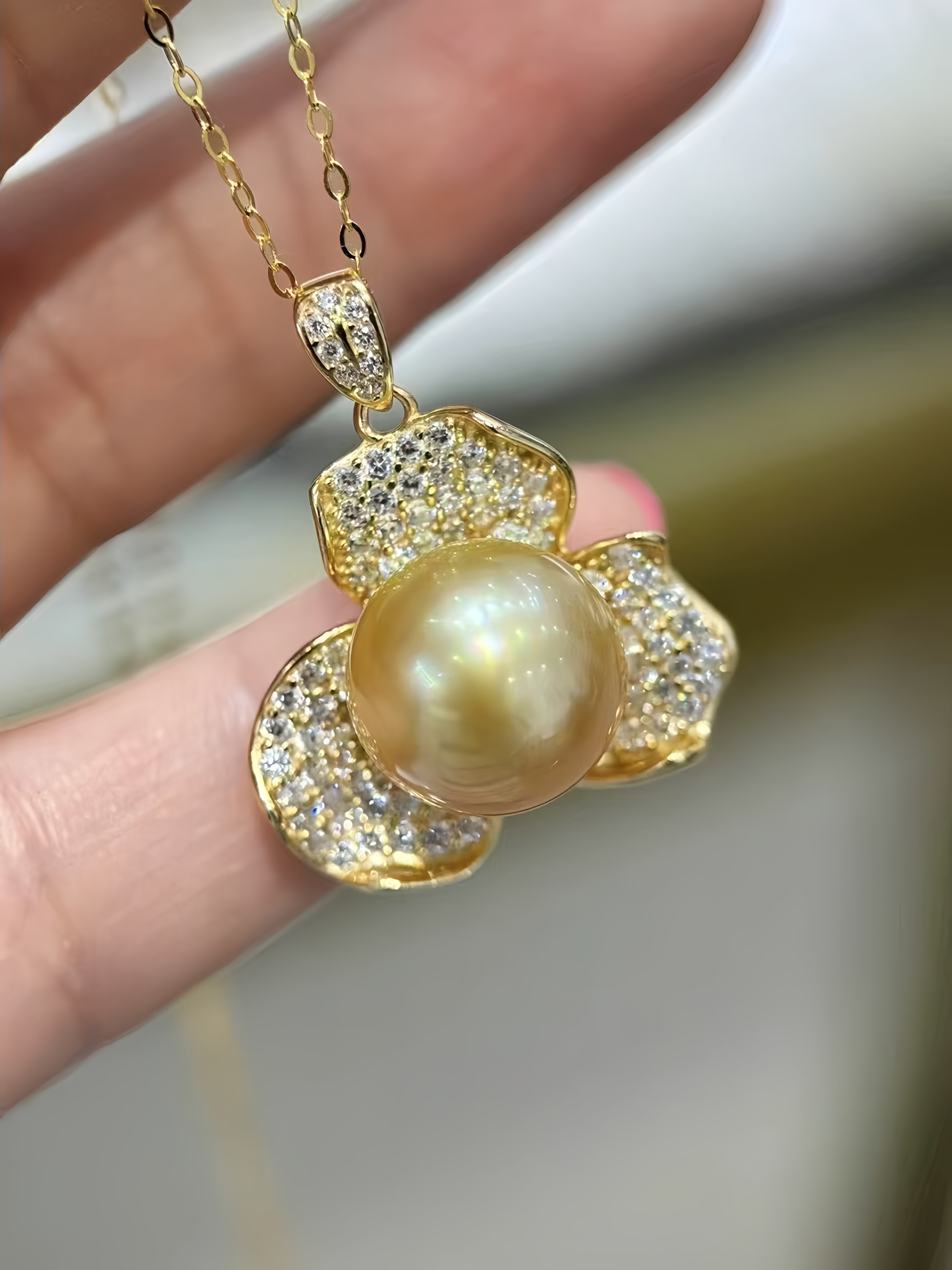 A gold pendant necklace with a large round pearl surrounded by pavé-set clear stones, held in a person's hand against a softly blurred light background.