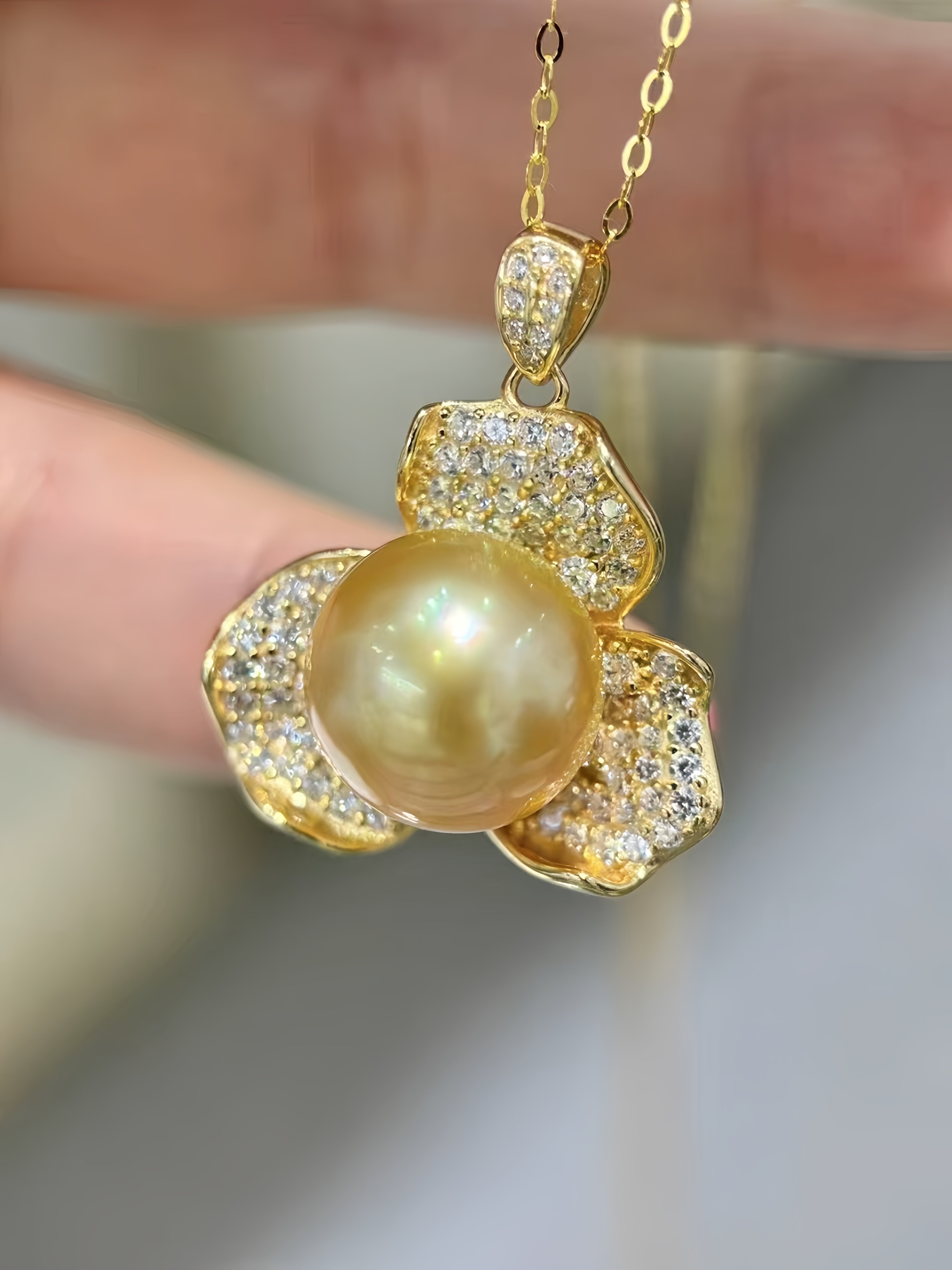 A gold flower-shaped pendant necklace featuring a large round pearl in the center and pavé-set clear stones on the petals, held by a hand against a soft blurred background.