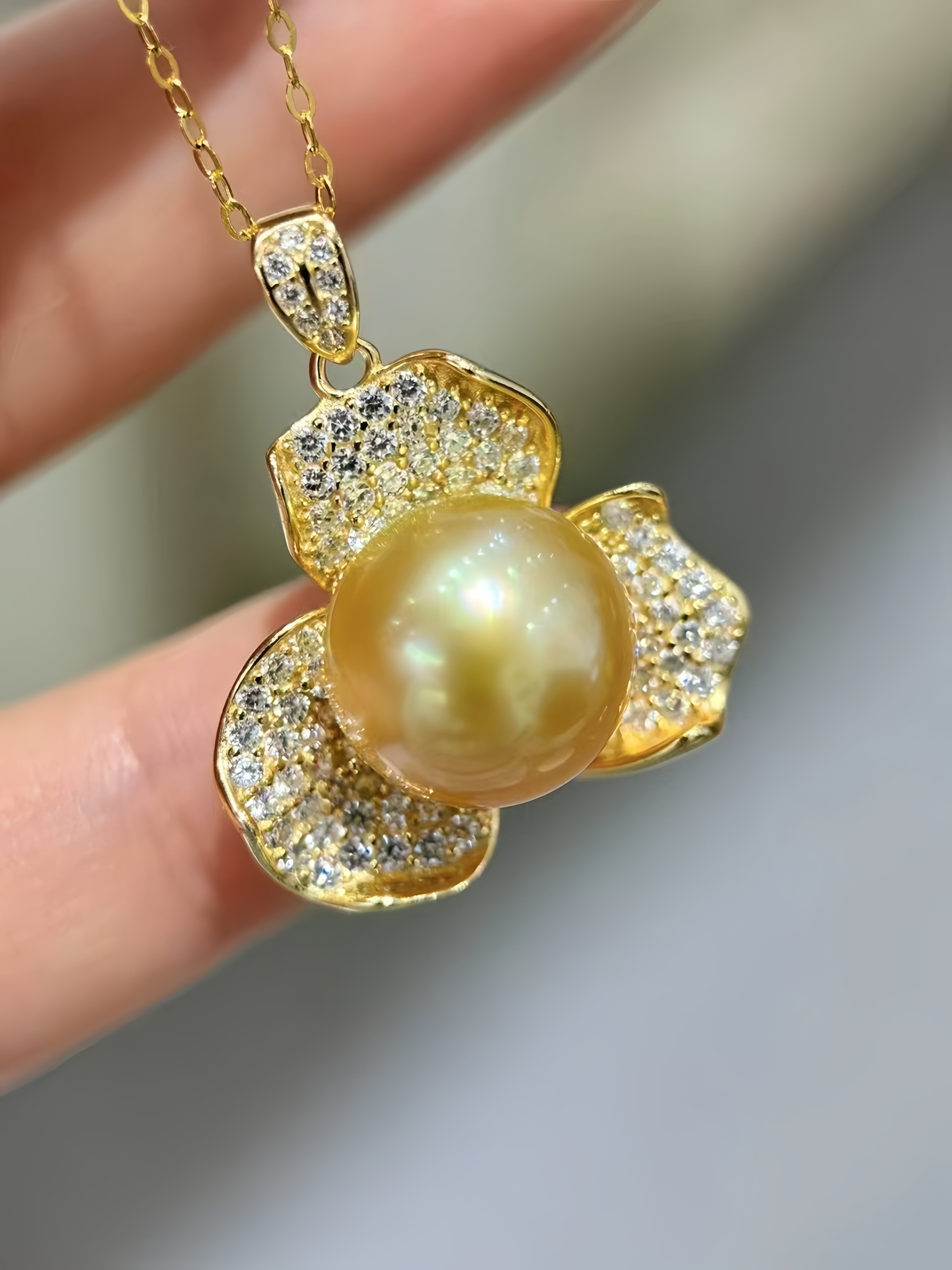 A gold pendant necklace featuring a large round yellow pearl surrounded by pavé-set clear stones in a flower-like shape, held between fingers against a soft gray background.