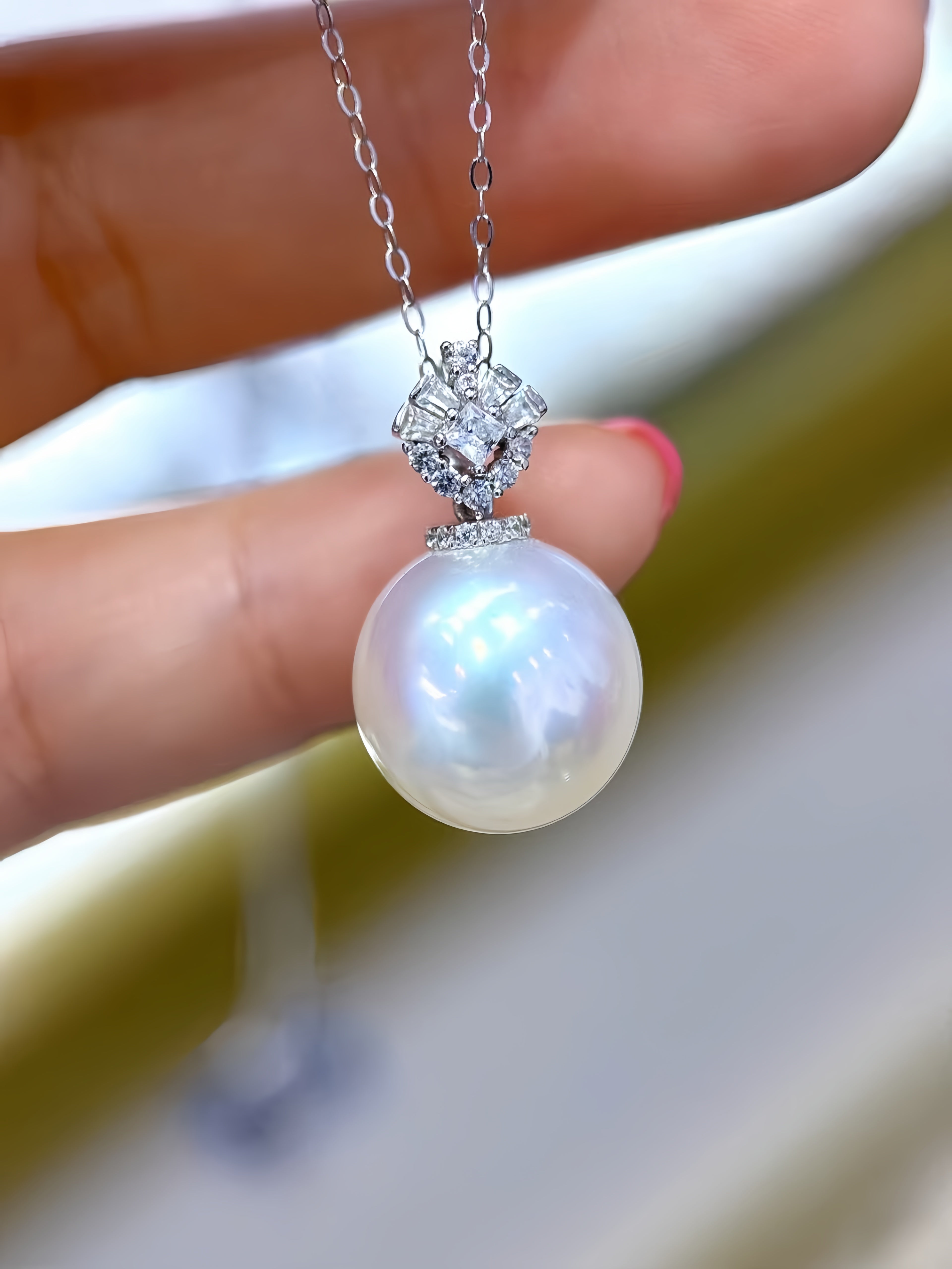 A close-up of a hand holding a silver pendant necklace featuring a large, lustrous white pearl and a sparkling diamond-like setting.