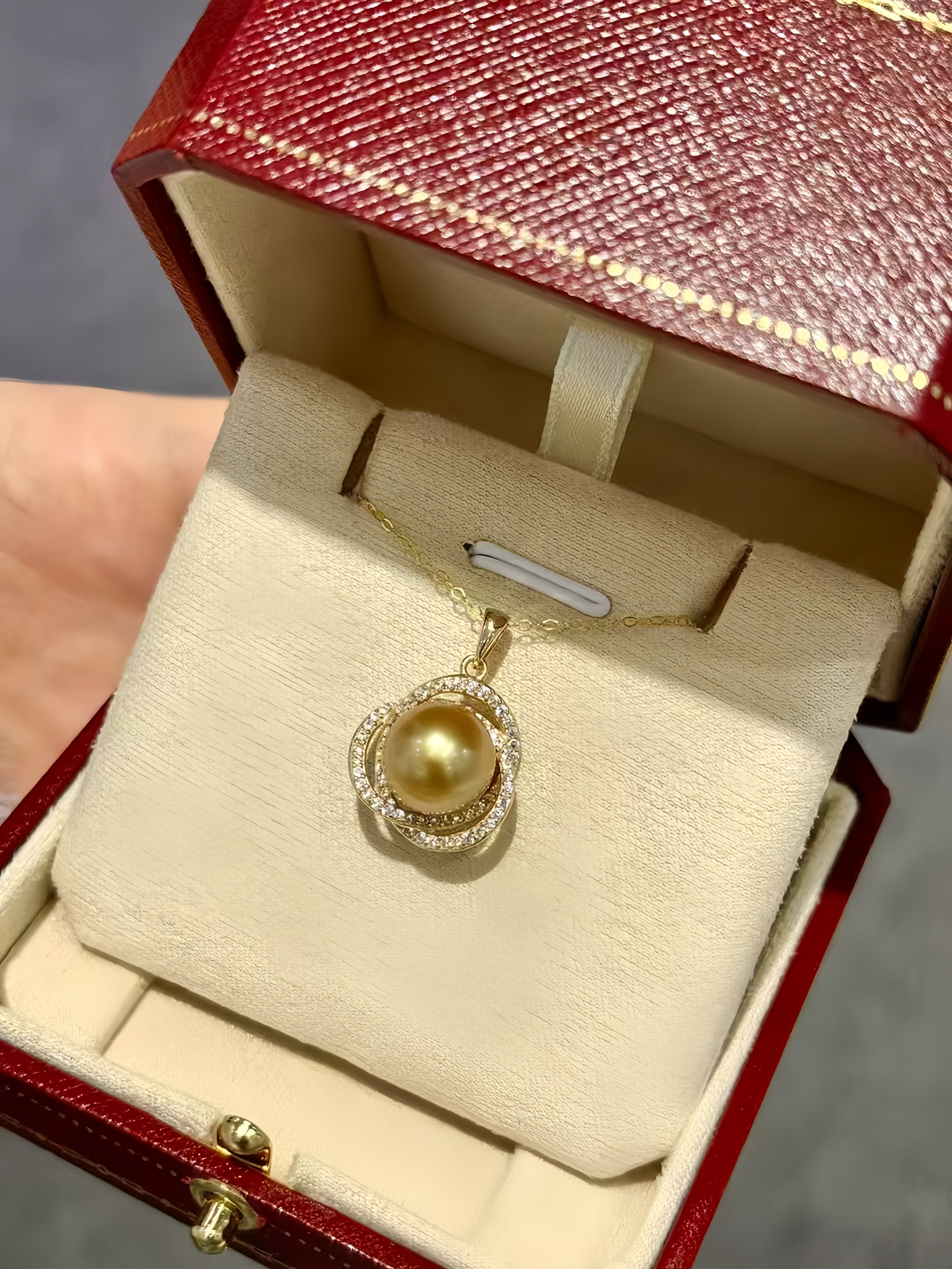 A gold pendant necklace with a large round yellow pearl and small clear stones, displayed inside an open red jewelry box with a cream velvet insert.