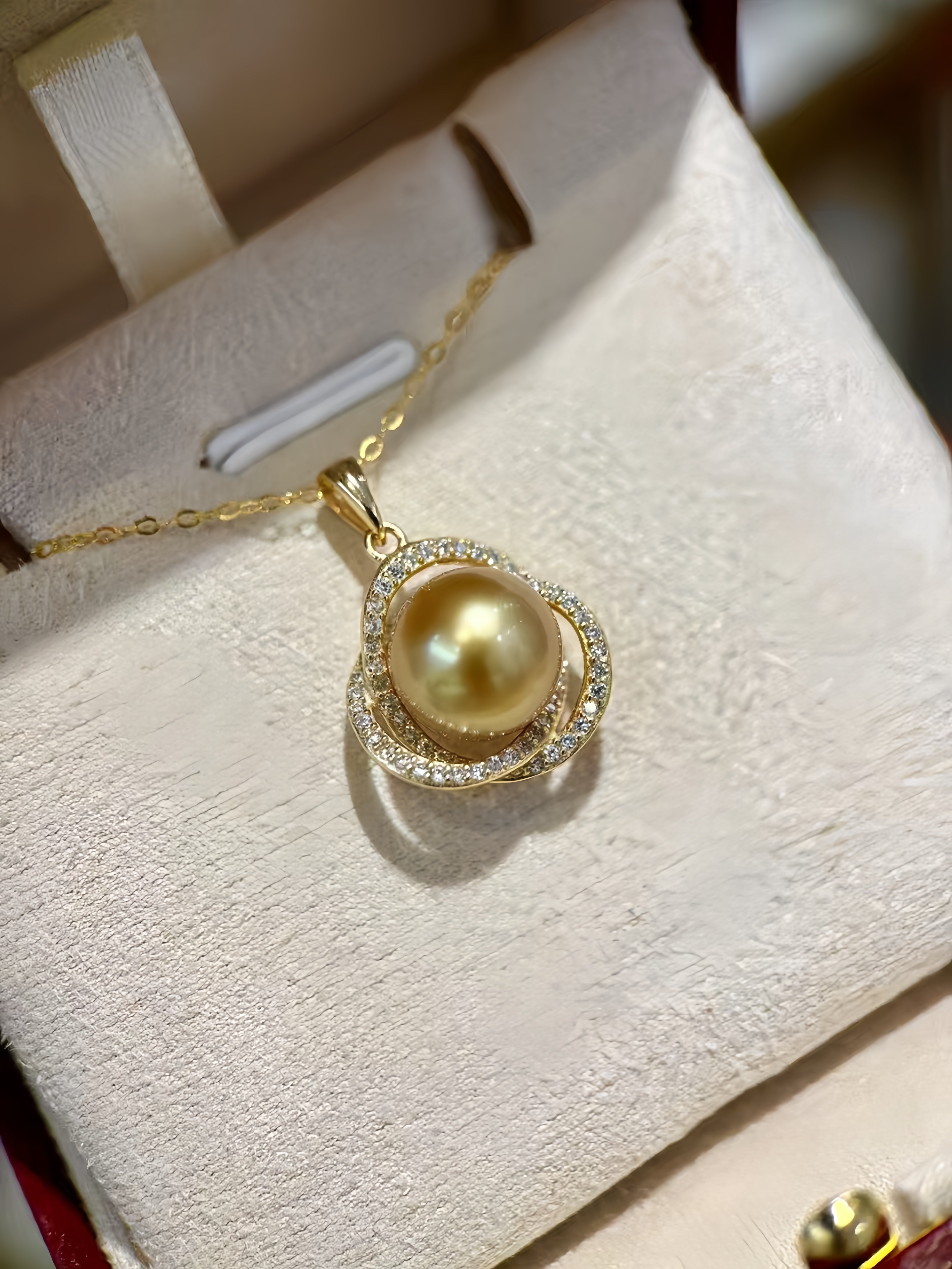 A gold pendant necklace featuring a large round yellow pearl surrounded by a halo of small clear stones, displayed on a cream-colored jewelry box.
