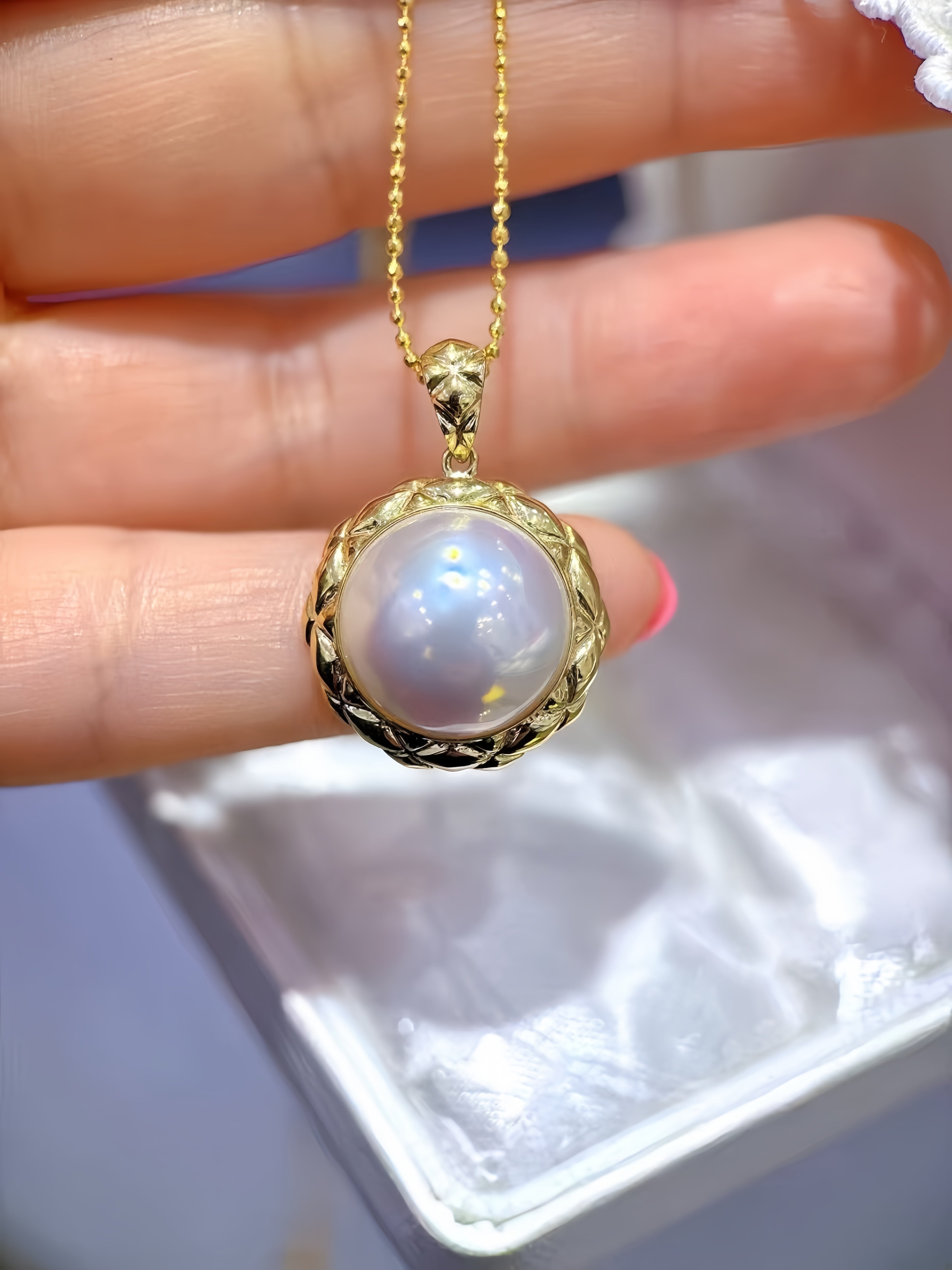 A gold pendant necklace with a large round iridescent pearl-like center and a textured gold bezel, held in a person's hand above a white jewelry box.