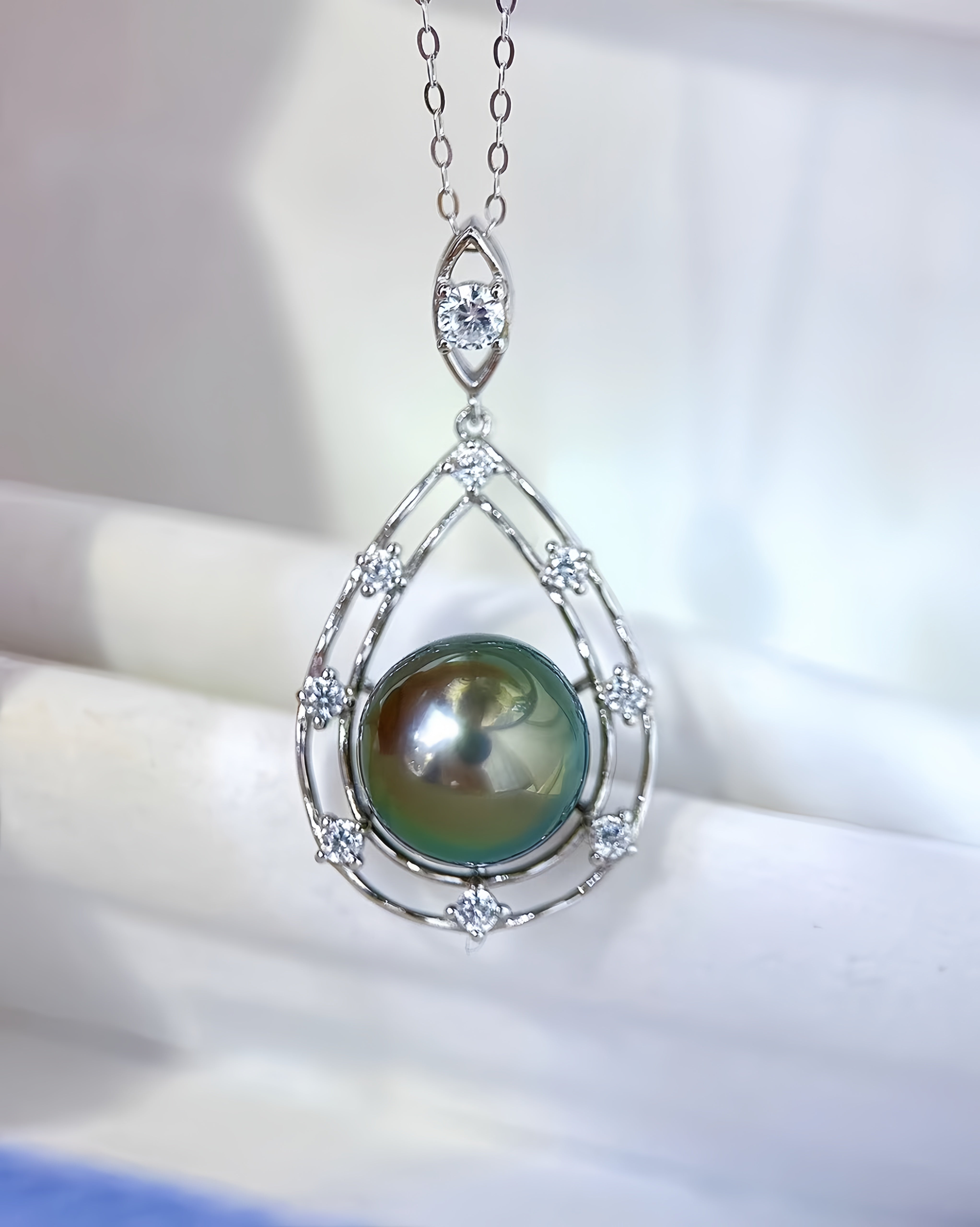 A silver teardrop pendant necklace featuring a large iridescent green pearl surrounded by small clear stones, hanging from a delicate silver chain.