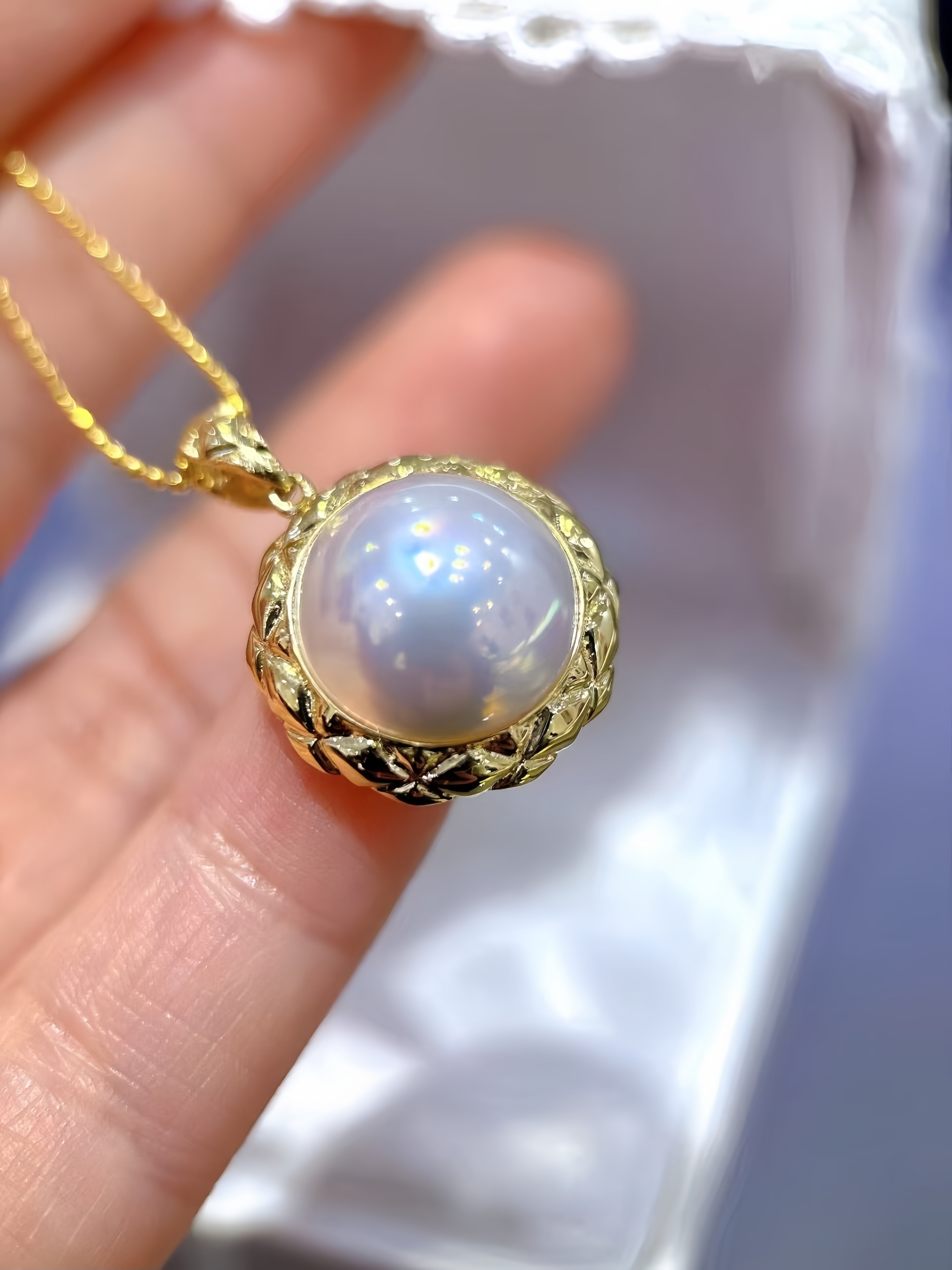 A gold pendant necklace with a round, lustrous white pearl-like center and an ornate textured gold bezel, held between fingers against a soft blurred background.