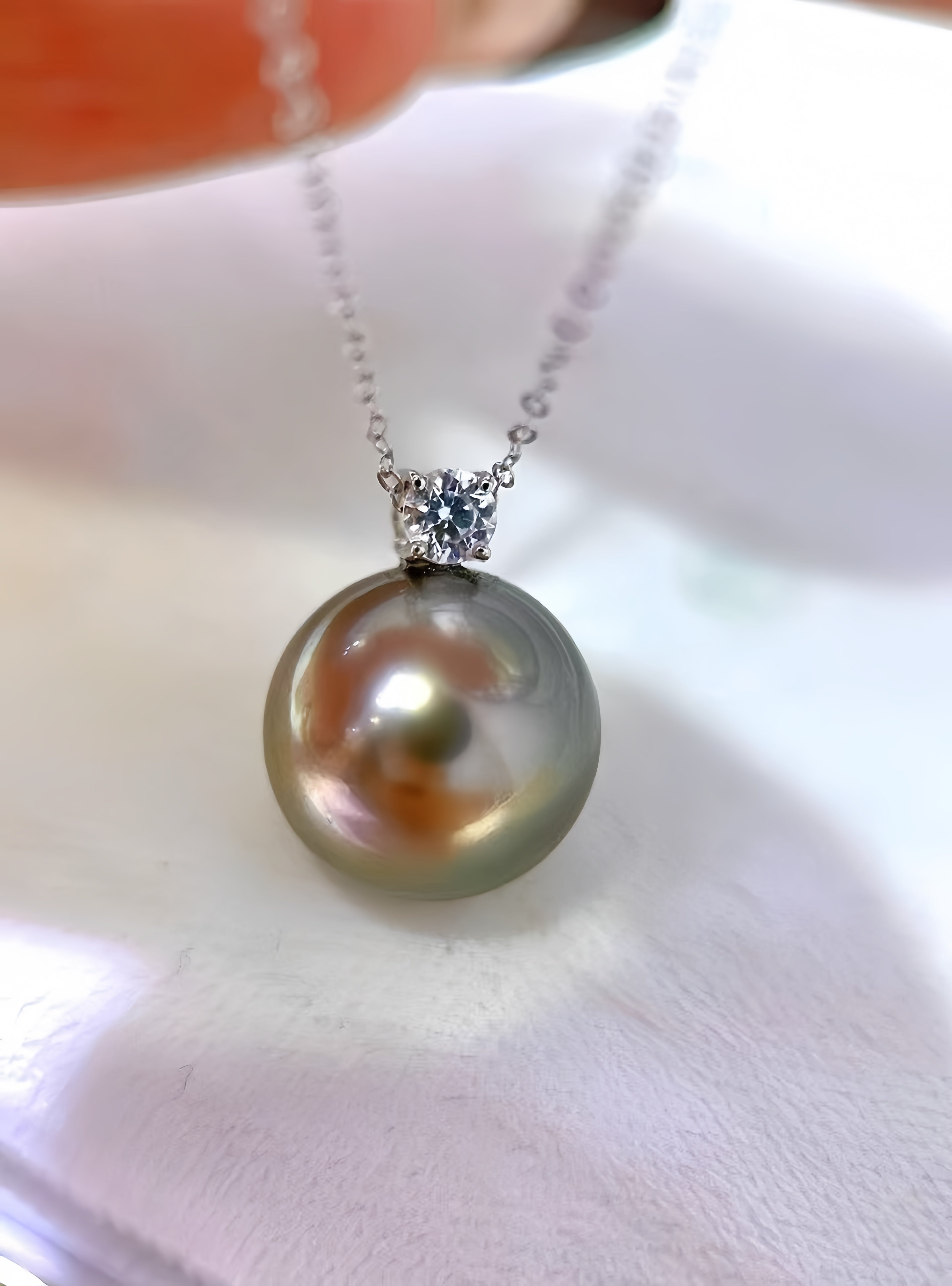 A close-up of a pearl pendant necklace on a delicate silver chain, resting on a soft white fabric surface. The pearl is iridescent with green, gold, and pink tones, set in a silver-toned bezel with a small clear gemstone.