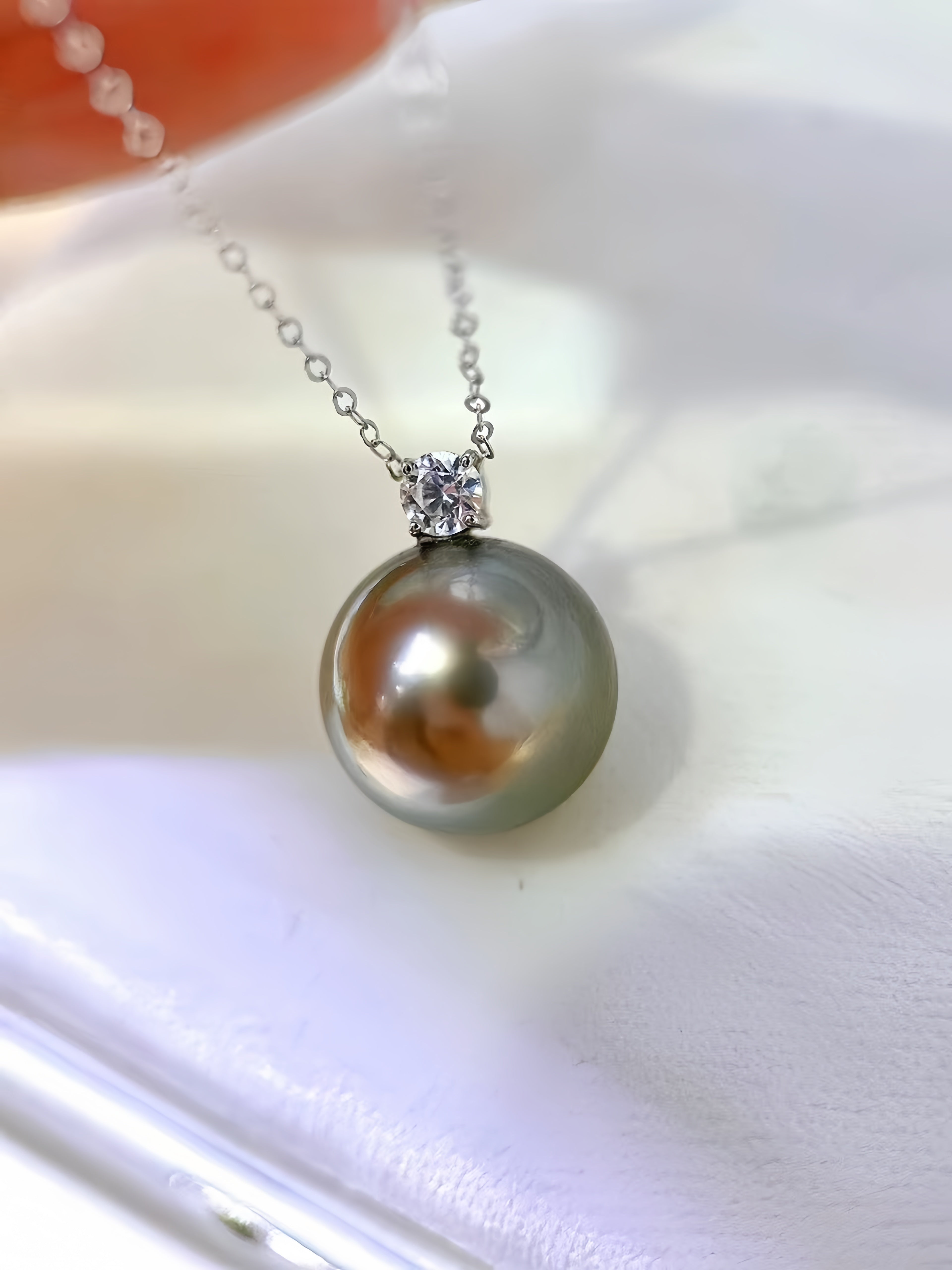 A close-up of a silver chain necklace with a round grayish-green pearl pendant and a small clear stone setting, resting on a white surface.
