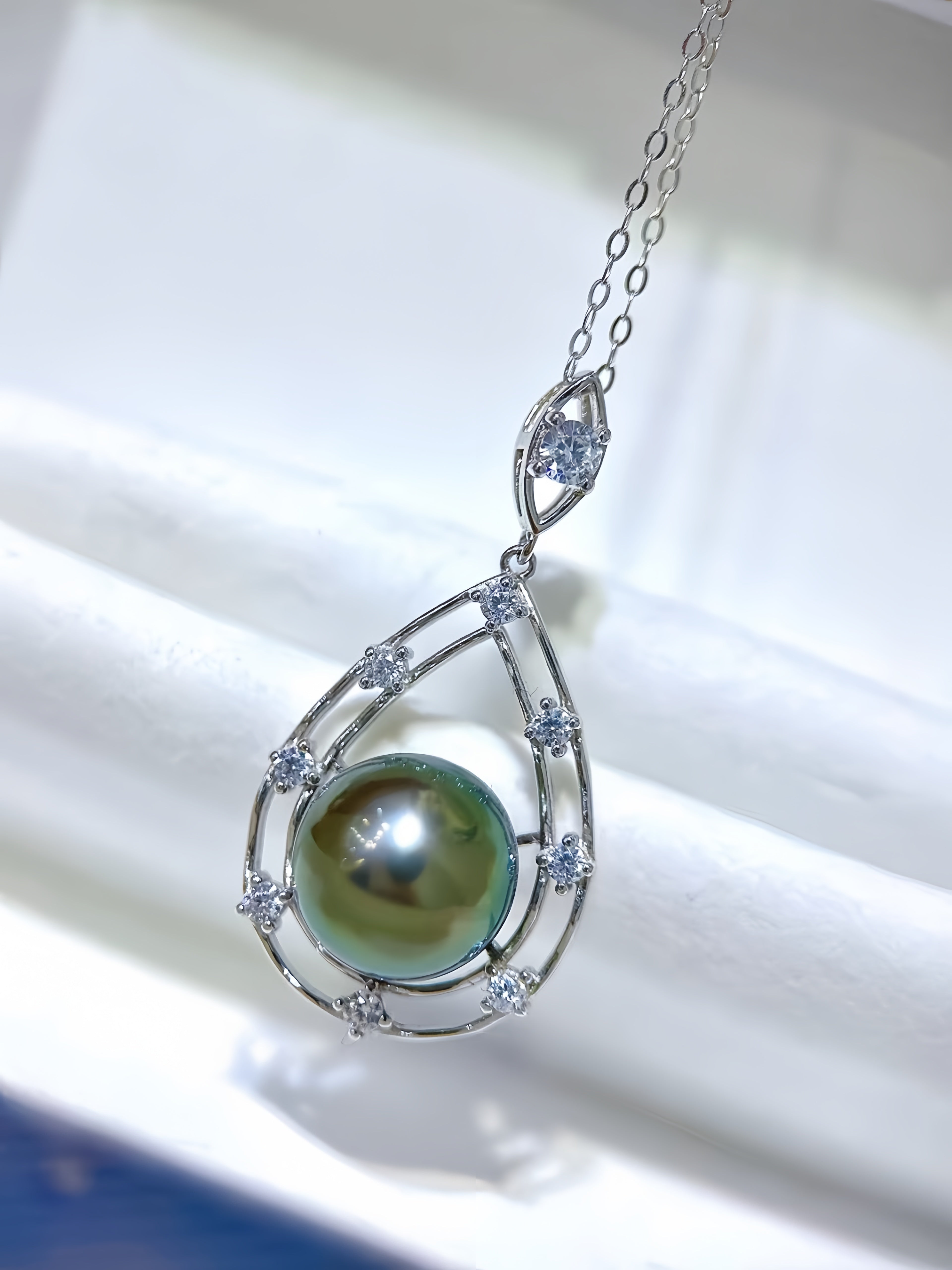 A silver teardrop pendant necklace featuring a large green-black pearl surrounded by small clear stones, displayed on a white jewelry box.