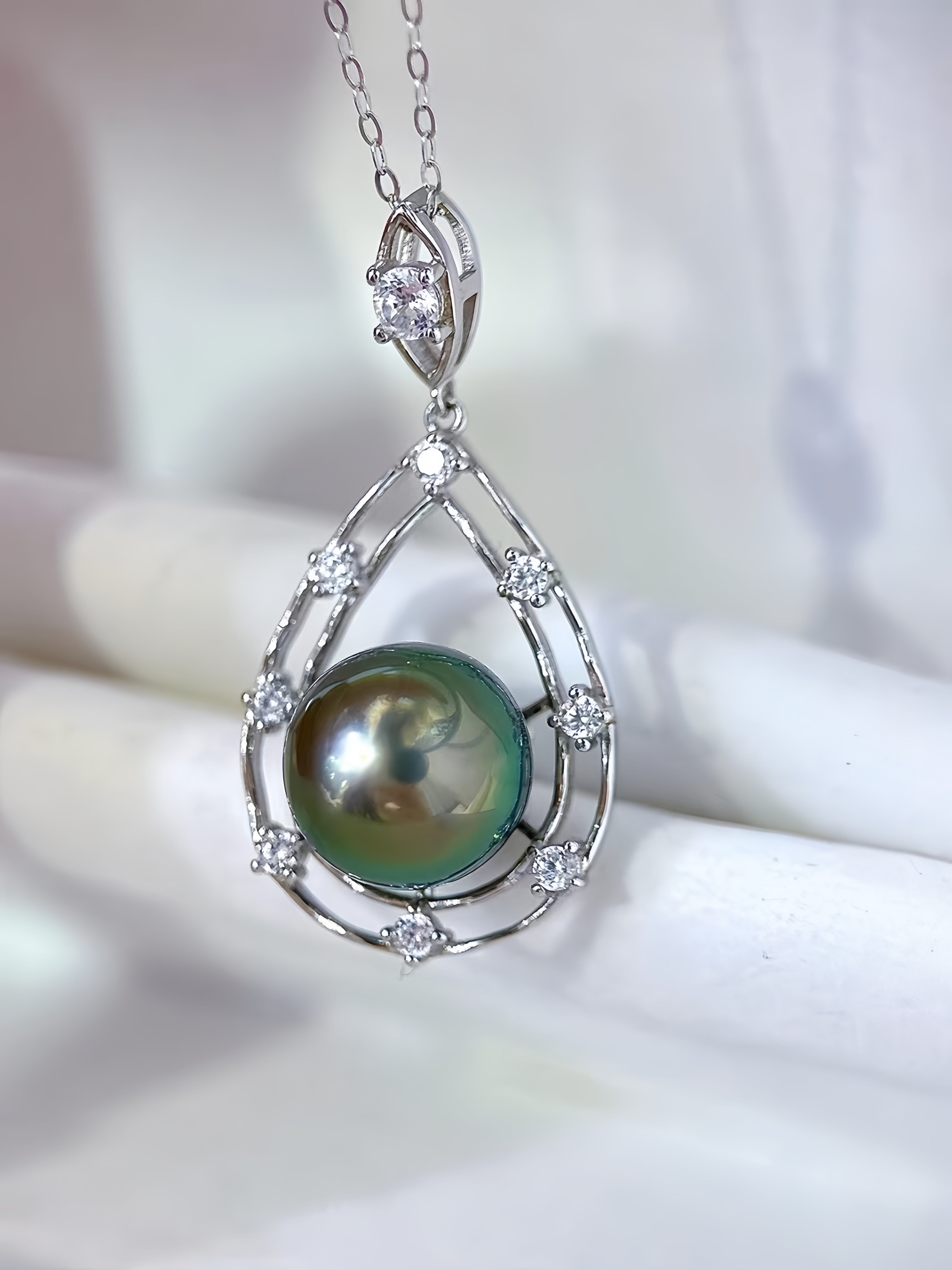 A silver teardrop pendant necklace featuring a round green iridescent pearl surrounded by small clear stones, hanging against a soft white background.