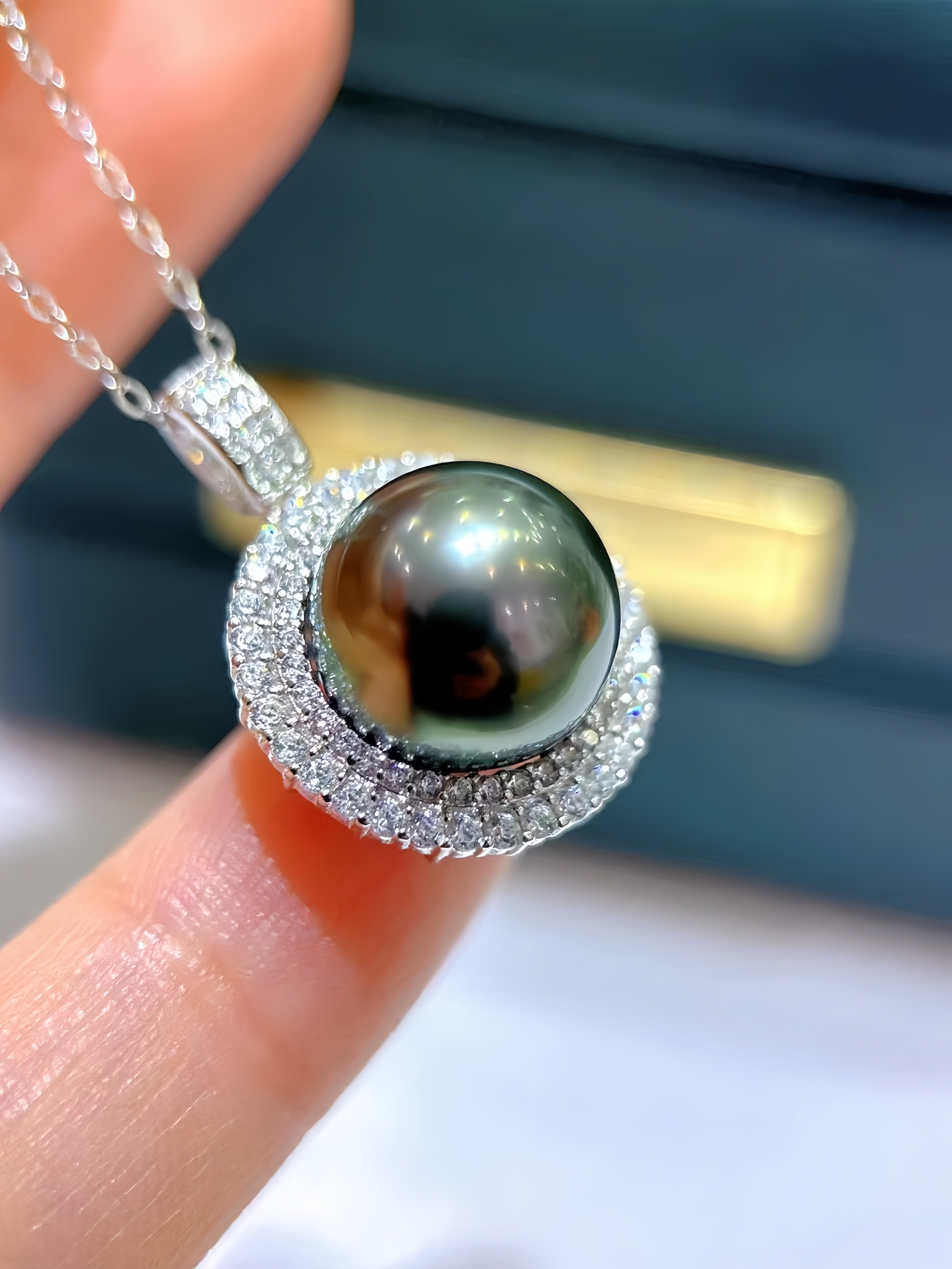 A close-up of a silver pendant necklace featuring a large, round black pearl surrounded by a halo of small clear stones, held between two fingers.