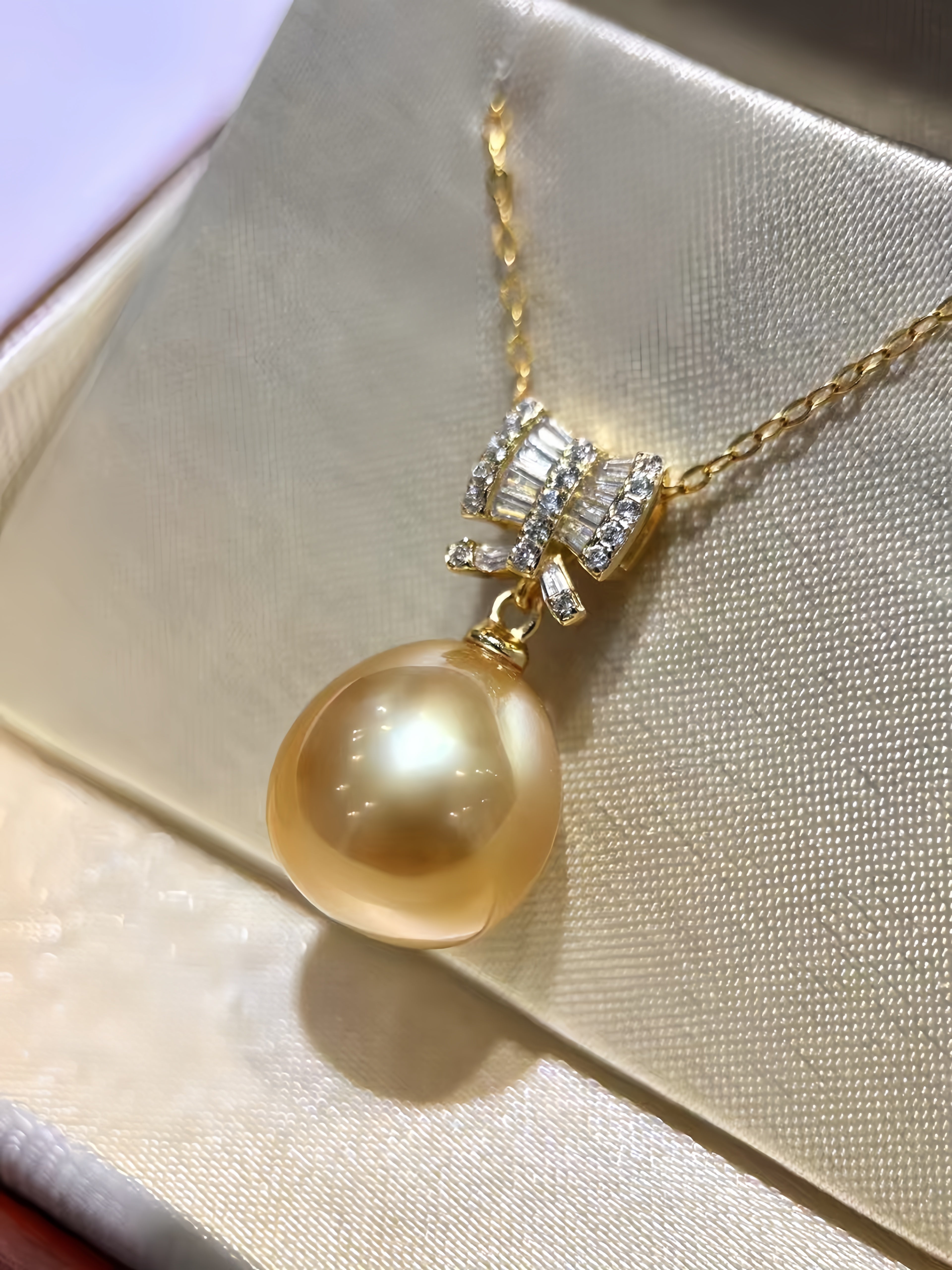 A gold pendant necklace featuring a large, lustrous round pearl pendant and a decorative bow-shaped setting with clear stones, displayed on a cream satin jewelry box.