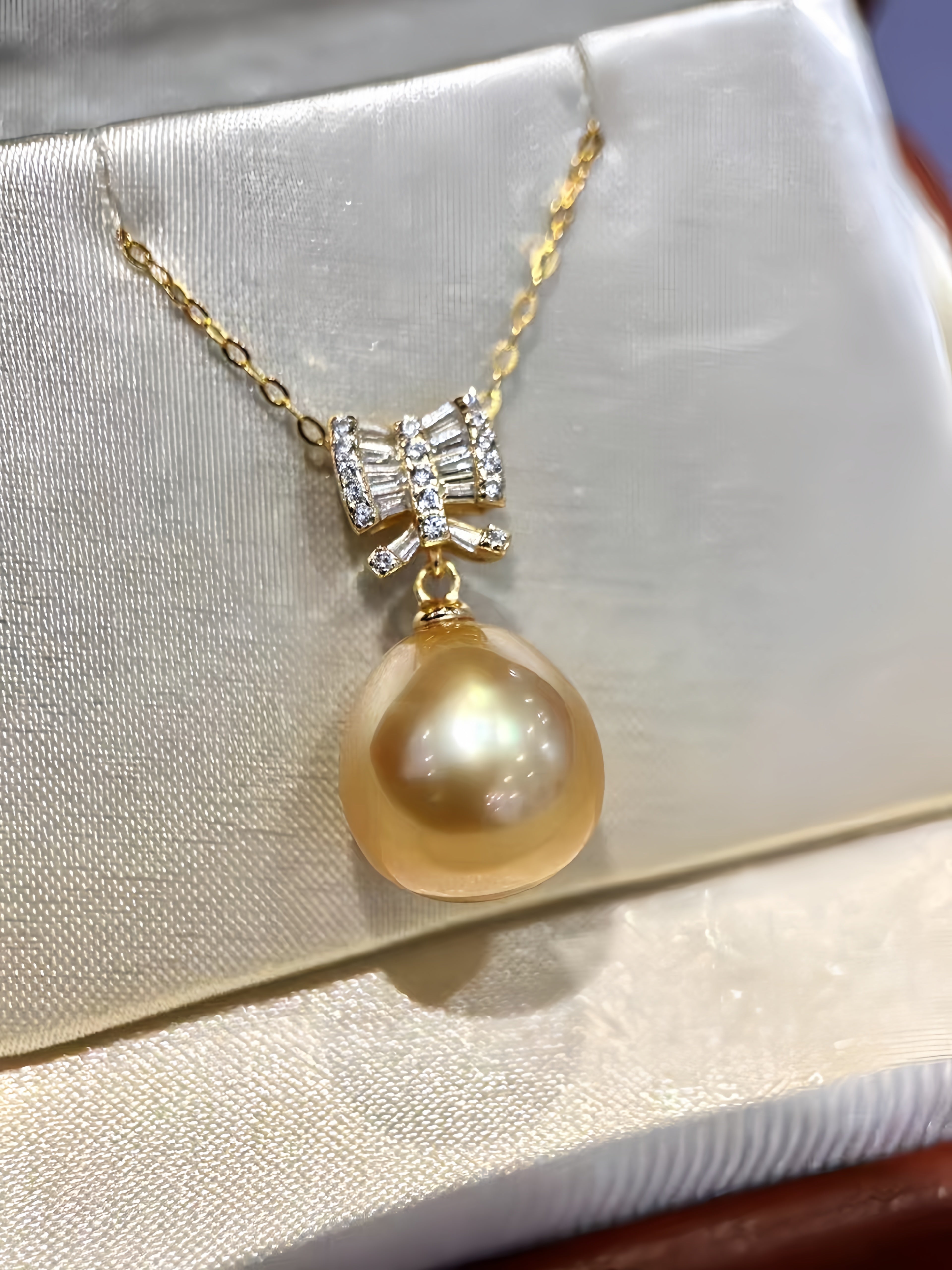 A gold pendant necklace featuring a large round champagne-colored pearl and a bow-shaped setting with clear stones, displayed on a cream satin jewelry box.