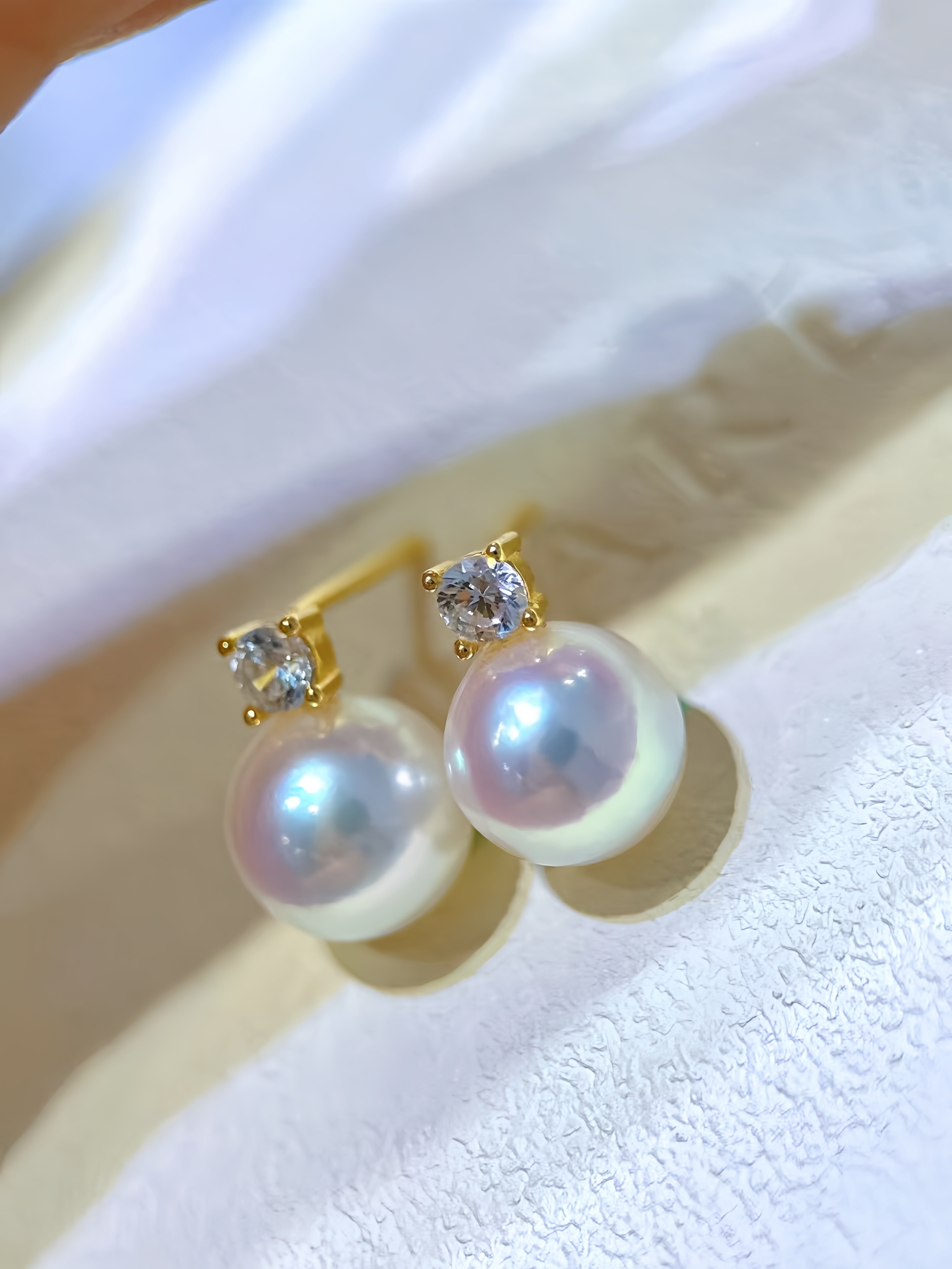 A pair of gold stud earrings with large iridescent white pearls and a small clear gemstone in each setting, displayed on a textured white surface.