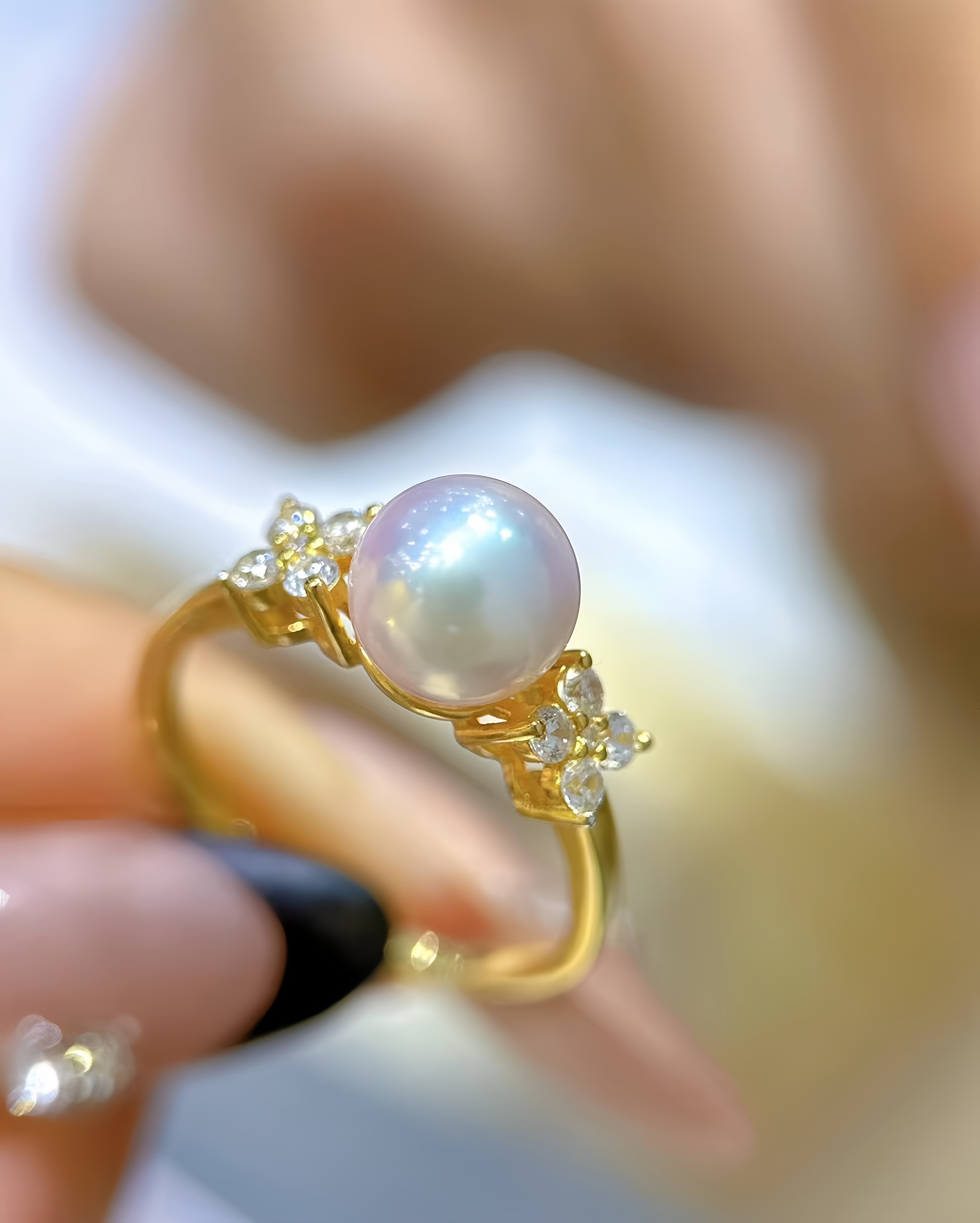 A gold ring featuring a large round iridescent pearl and small clear accent stones, held by a hand with manicured nails.