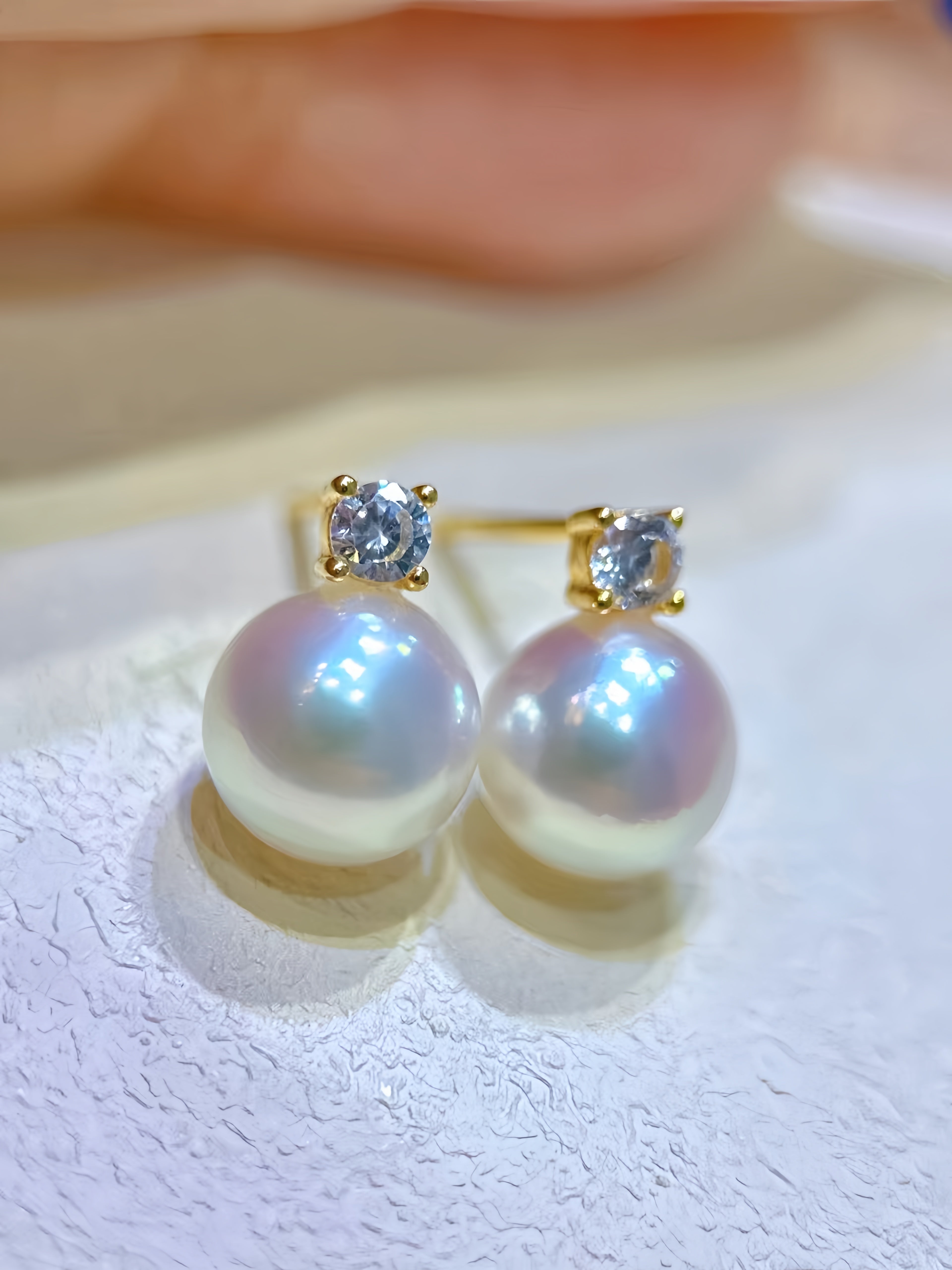 A pair of gold stud earrings with round white pearls and a small clear gemstone in a prong setting, resting on a textured white surface.