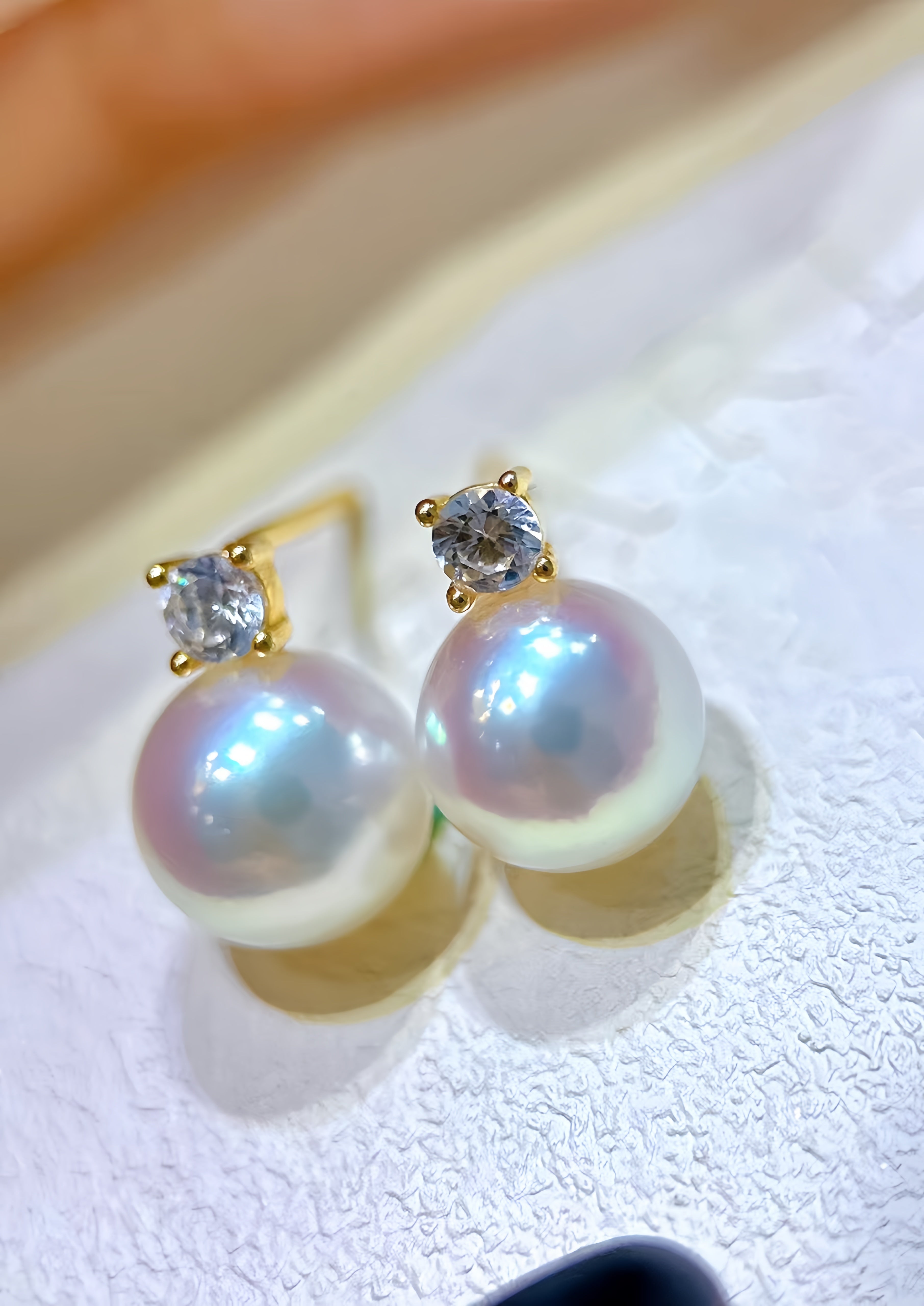 A pair of gold stud earrings featuring large, lustrous iridescent freshwater pearls and a small clear round-cut stone in a four-prong setting above each pearl, displayed on a textured white surface.