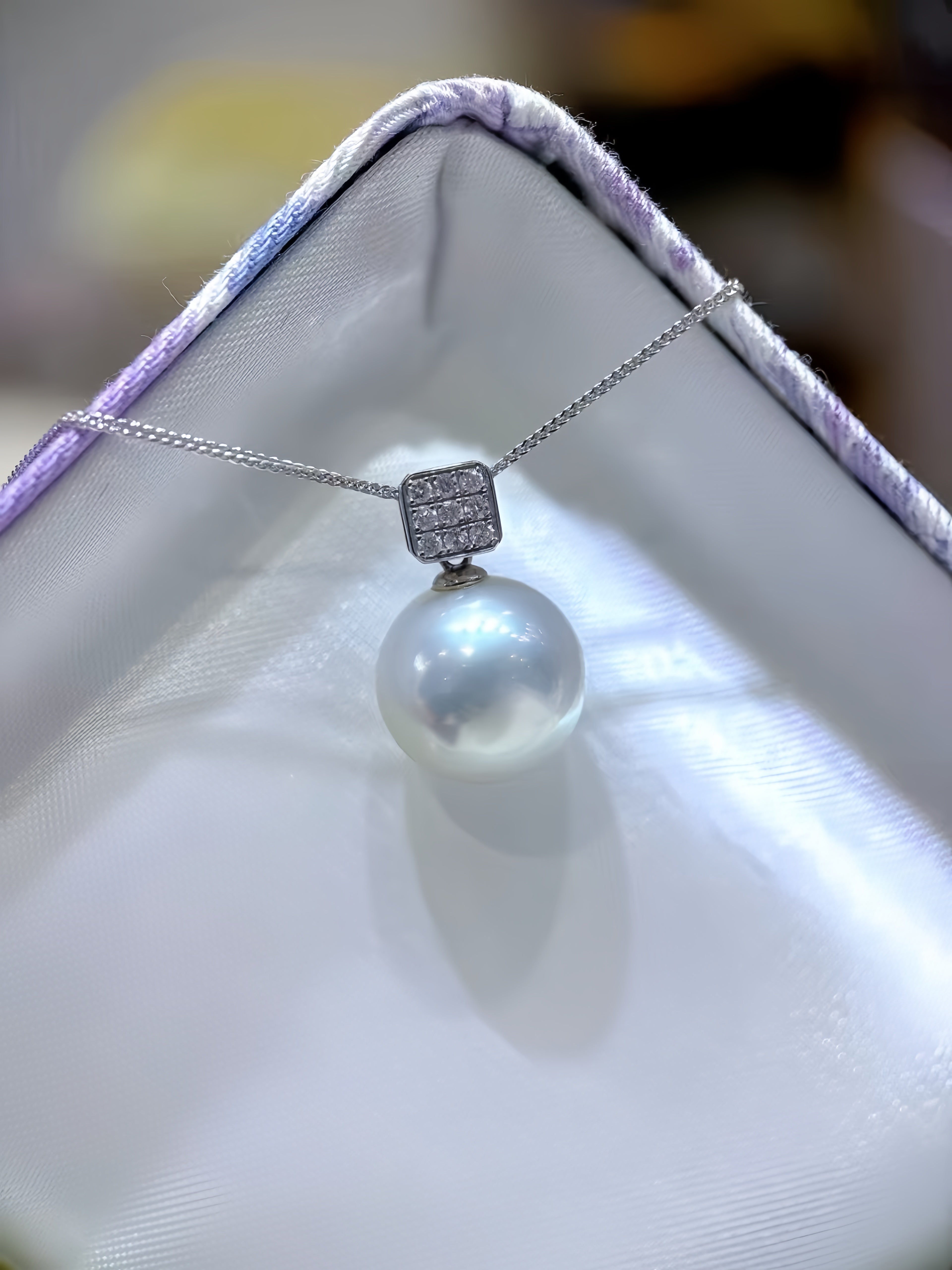 A silver pendant necklace featuring a single lustrous white pearl and a square pavé-set stone cluster, displayed on a white jewelry box.