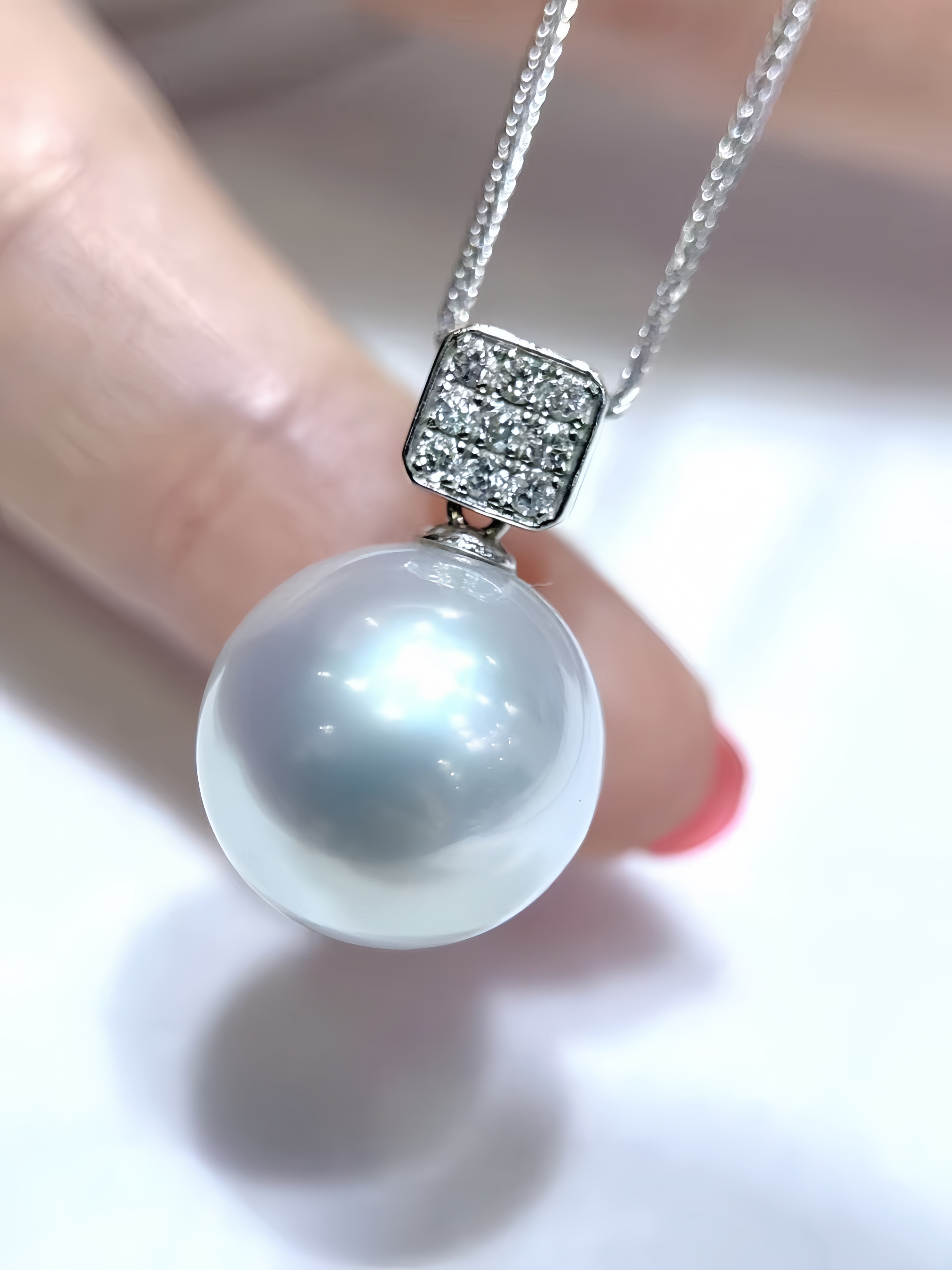 A close-up of a pearl pendant necklace held by fingers. The pendant features a large, lustrous white pearl suspended from a silver-toned setting with small clear stones, on a delicate silver chain.