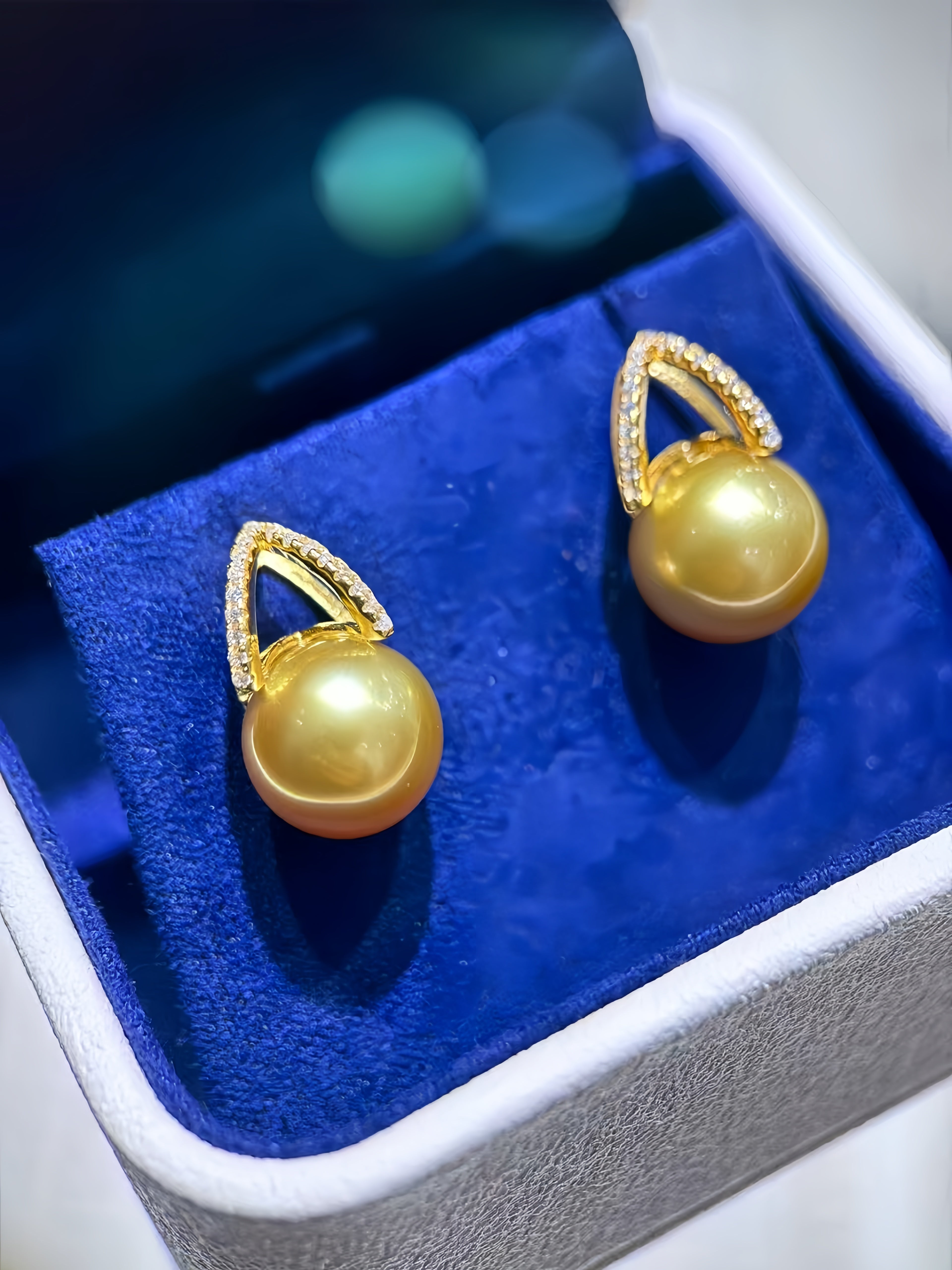 A pair of gold drop earrings with round yellow pearl drops, displayed in a white jewelry box with a blue velvet insert.