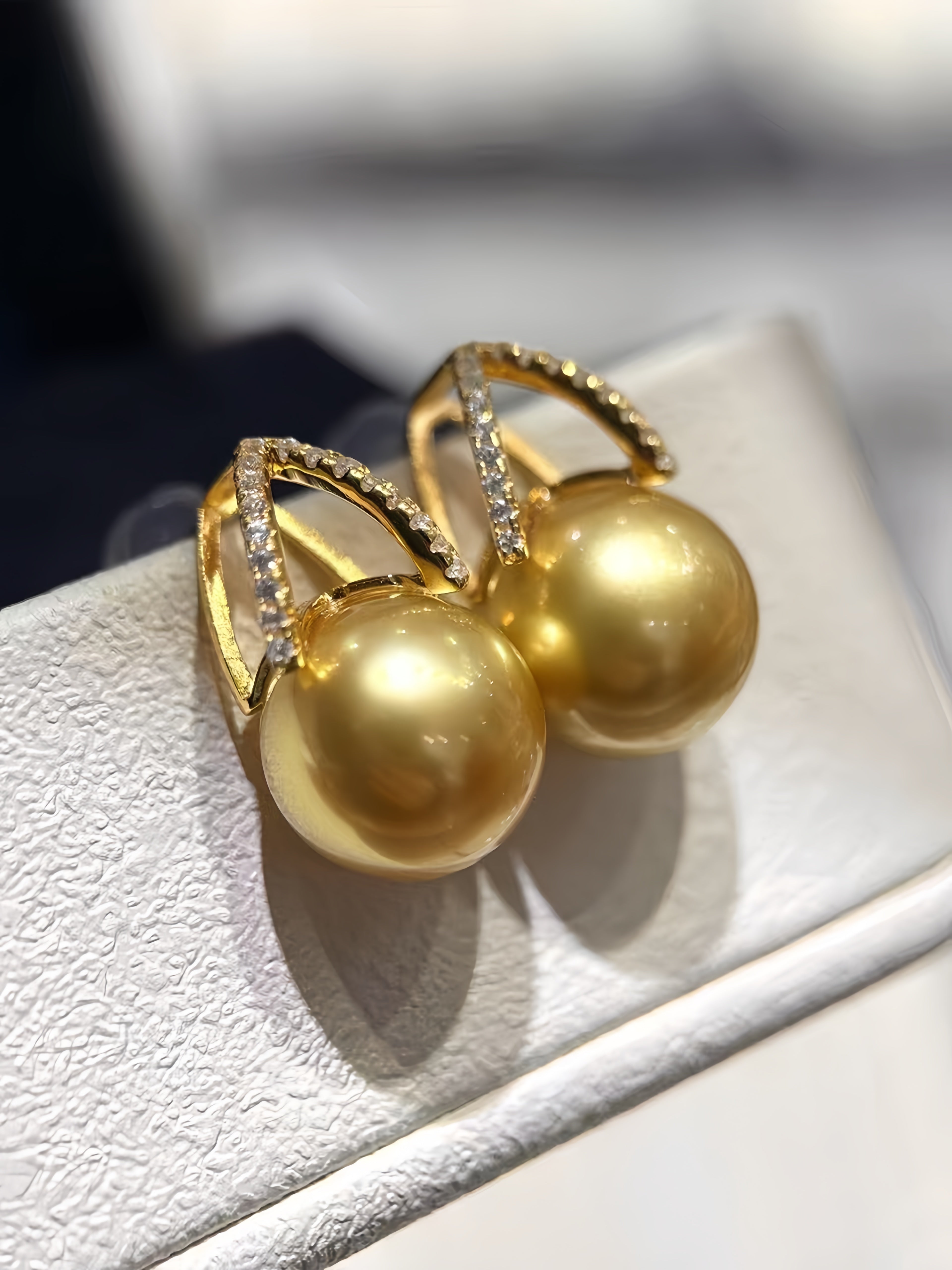 A pair of gold hoop earrings with large round pearl-like drops and small clear stones along the hoops, displayed on a white jewelry stand.