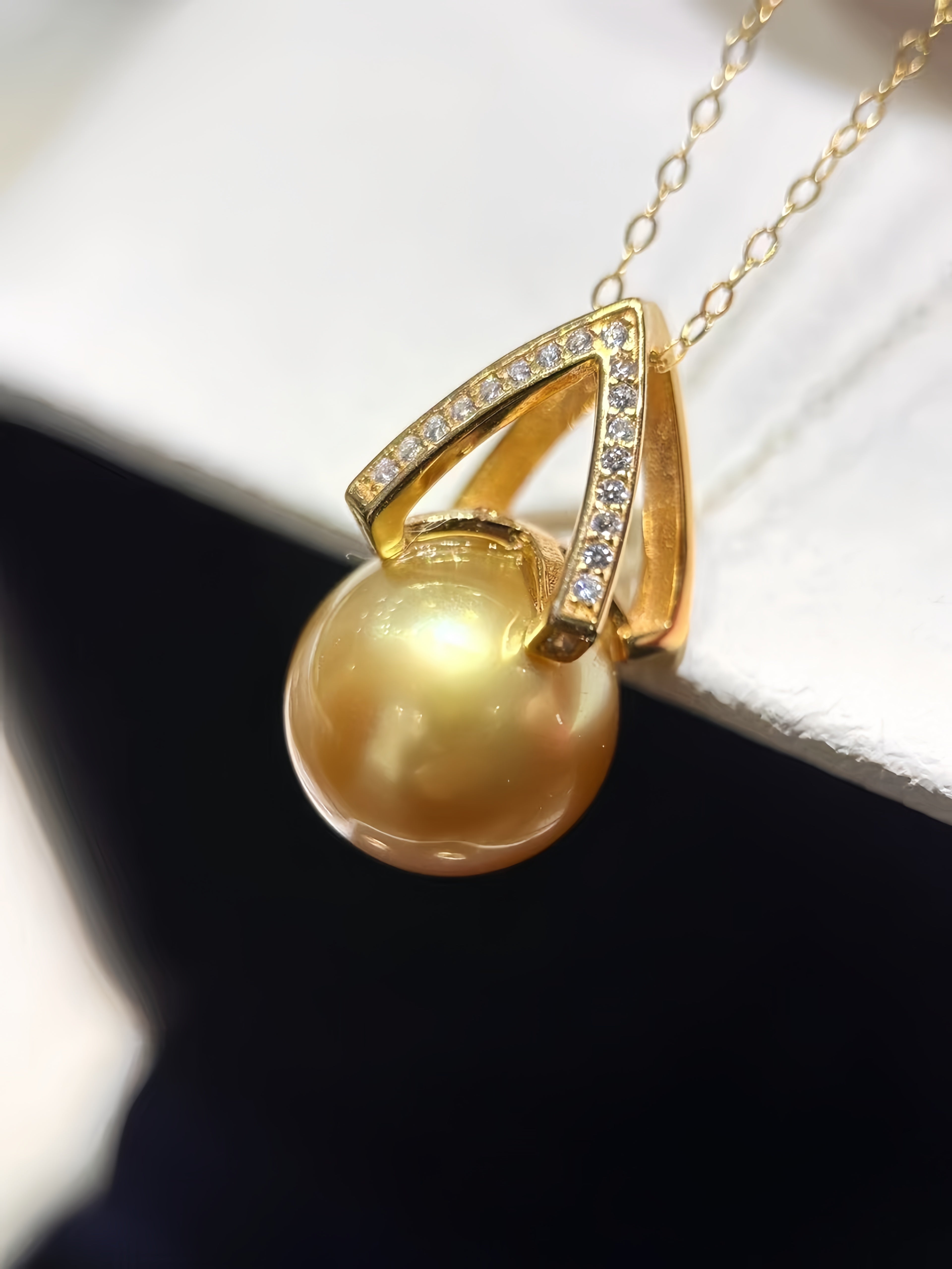 A gold pendant necklace featuring a large, lustrous golden pearl suspended beneath a geometric gold frame adorned with small clear stones, shown against a white and black background.