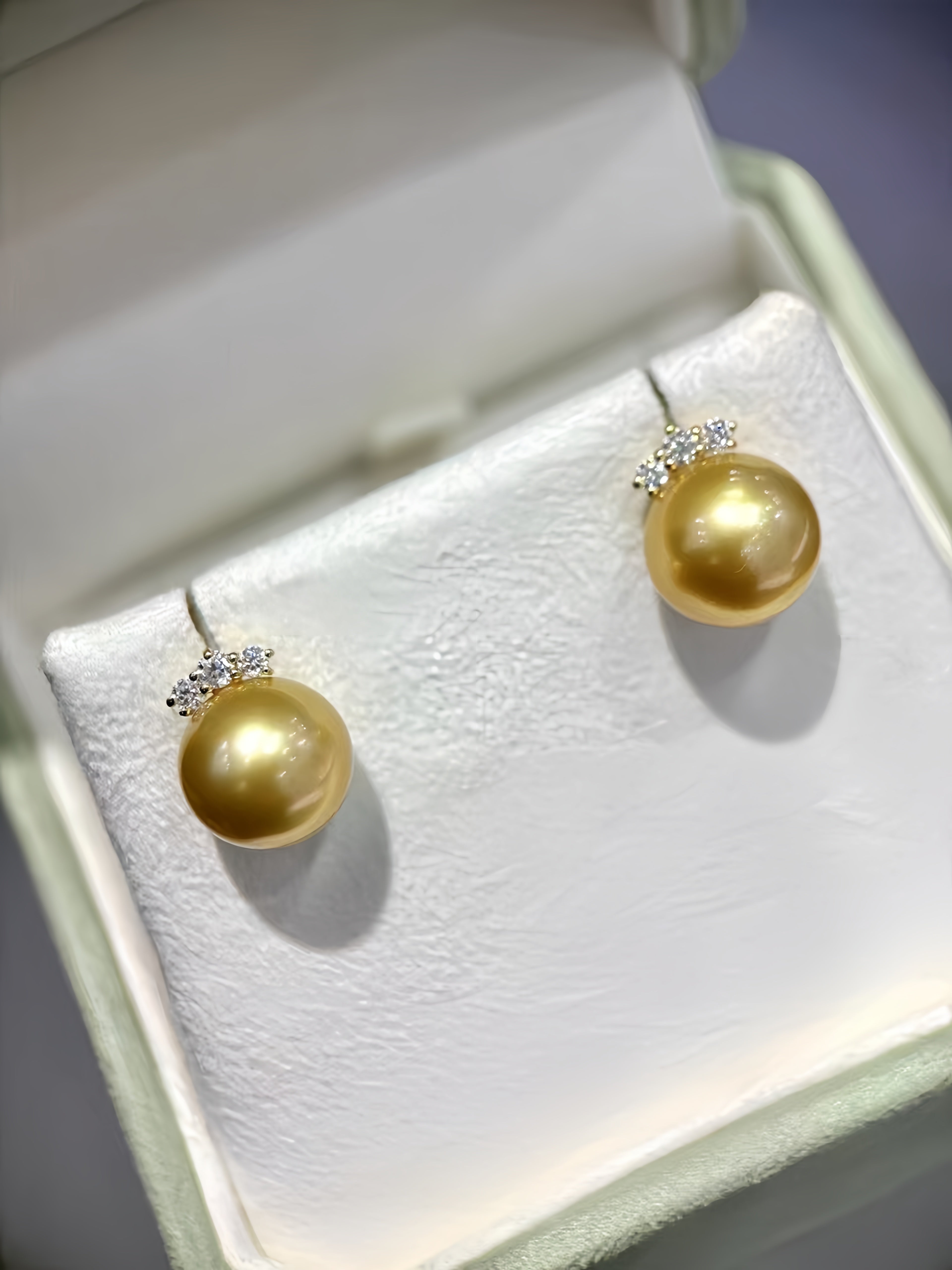 A pair of gold pearl stud earrings with small clear crystal accents, displayed on a white textured jewelry box insert.