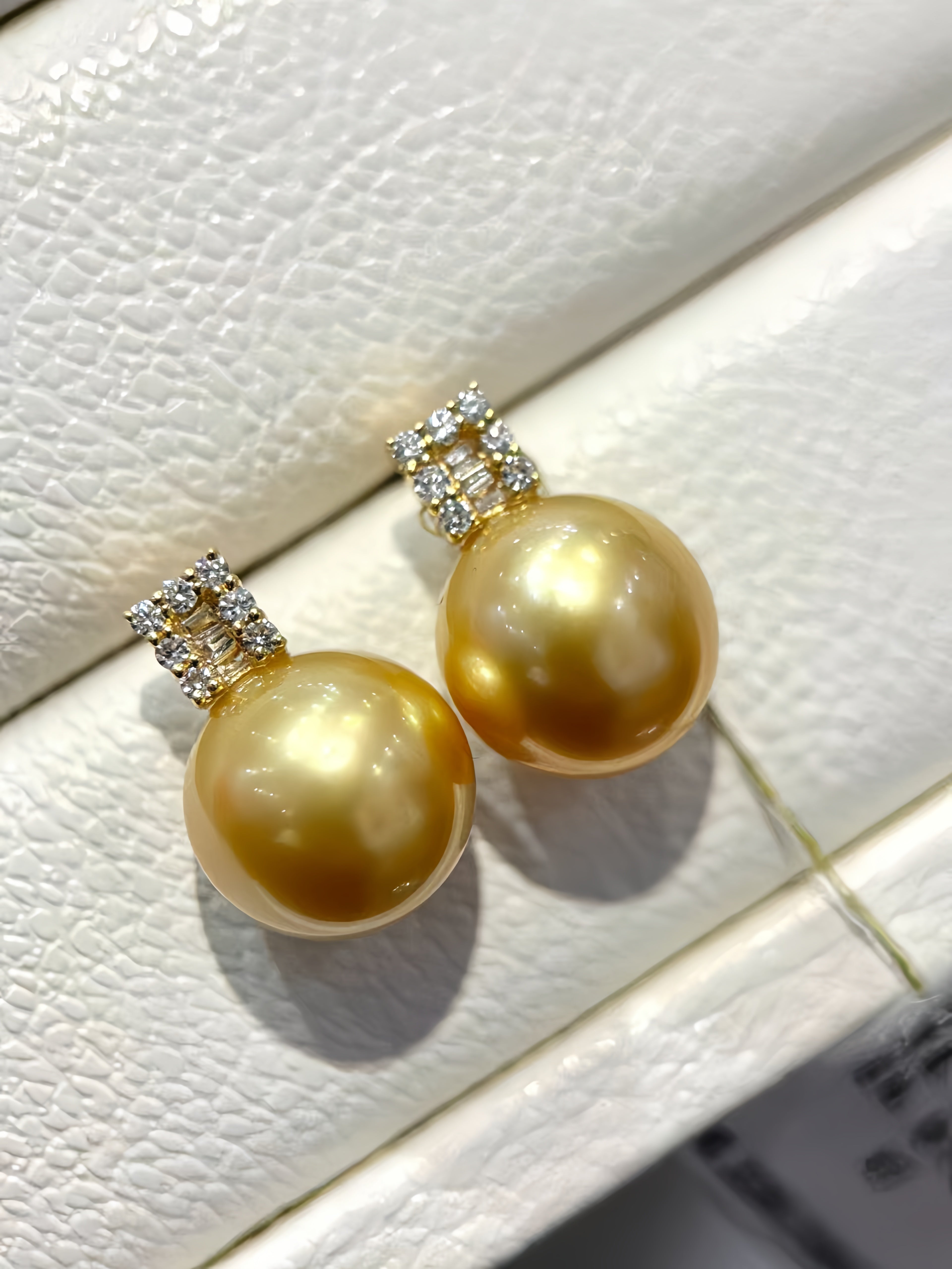 A pair of gold-toned drop earrings with large round freshwater pearls and small clear crystal clusters at the top, displayed on a textured white jewelry box.